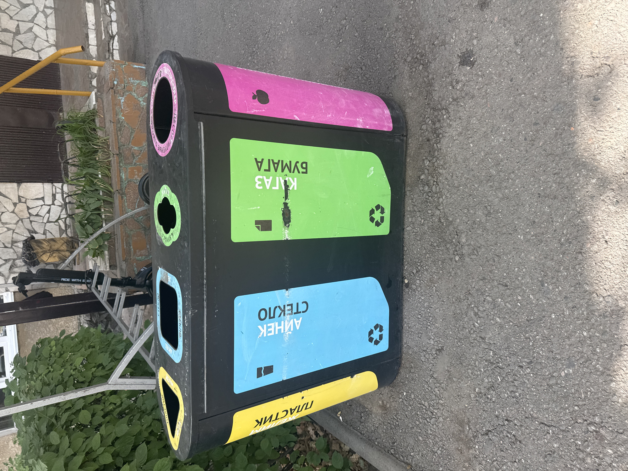 An outdoor, eye-level, full shot captures a multi-compartment recycling bin positioned on an asphalt surface. The bin is primarily black with brightly colored labels and openings. From left to right, the compartments are designated by a yellow label with Cyrillic text, a blue label with Cyrillic text and a recycling symbol, a green label with Cyrillic text and a paper icon, and a pink section with an apple icon. The yellow label reads "ПЛАСТИК" (PLASTIC). The blue label reads "СТЕКЛО" (GLASS). The green label reads "БУМАГА" (PAPER) and the icon appears to be a folded piece of paper. The pink section may represent organic waste or another category. Behind the recycling bin, a building with a stone facade and a brown door is visible, along with green foliage and a metal structure that resembles a bicycle rack. A black electric scooter with "RIDE WITH EVER" branding is also present. The scene appears to be located in Bishkek City, Kyrgyzstan, possibly near an entrance to a residential building or commercial establishment, given the presence of the building facade and the public amenity of a recycling bin. The lighting suggests it is daytime.