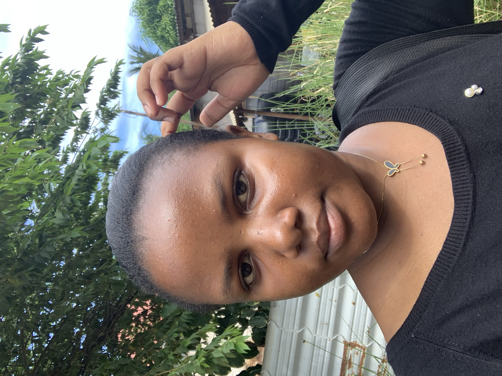 The image presents a close-up outdoor portrait of a dark-skinned female individual, approximately in her late teens to early thirties, looking directly at the camera with a neutral expression. Her short, dark hair is closely cropped. Her complexion appears shiny, exhibiting small, scattered epidermal irregularities across the forehead and cheeks. She wears a black long-sleeved top with a ribbed crew neck. Around her neck is a gold-colored chain necklace featuring a black and gold butterfly pendant with two small dangling gold beads. A decorative pin, appearing to be composed of small pearls and clear stones, is affixed to her top on the left chest area. A black strap from a backpack is visible over her right shoulder.
Her right hand is raised to the right side of her head, with the index finger gently touching the hair near her temple; the remaining fingers are partially curled.
The background depicts a residential outdoor setting. To the left, dense green foliage from a large tree dominates the upper portion, with a light-colored, possibly corrugated metal fence showing signs of discoloration or minor rust below it. To the right, a bright sky with scattered white clouds is visible above more green foliage, including what appear to be palm fronds, and a utility pole. Lower down on the right, tall green grass or weeds are prominent, behind which parts of a building with a corrugated metal roof are discernable. The environmental features, including the lush vegetation, utility infrastructure, and construction style, are consistent with a residential area within the city of Dar es-Salaam, Tanzania.