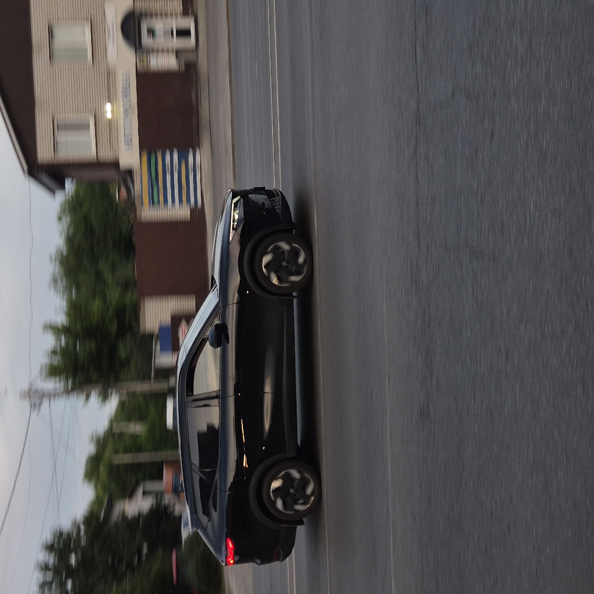 A black crossover SUV is driving on a paved road in a city. The car is dark and sleek, with its headlights and taillights illuminated. The wheels are visible and appear to be spinning, indicating motion. The background shows buildings and trees, suggesting an urban or suburban environment. The sky is overcast. A sign on one of the buildings appears to be in Russian. The overall scene suggests a snapshot of everyday life in a Russian city. The image appears to have been taken from a moving vehicle, as indicated by the motion blur in the background and the way the car is captured in profile.
