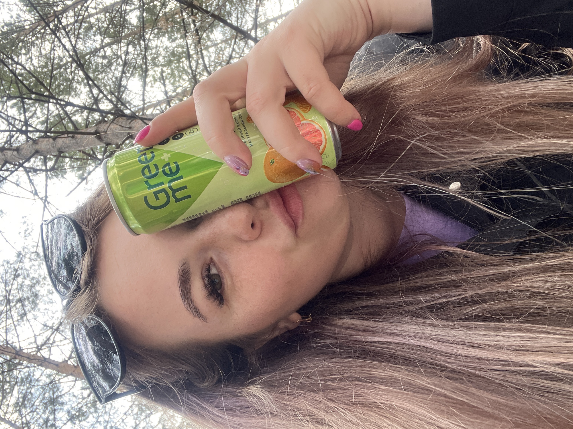 A young woman with long, light brown hair is holding a can of Green Me grapefruit-flavored beverage in front of her face. She has sunglasses perched on her forehead and is wearing a light purple shirt under a dark jacket. Her nails are painted; some with pink polish and others with a marbled design. The background shows tree branches against a bright sky. The location is Chaikovsky, Russia.
