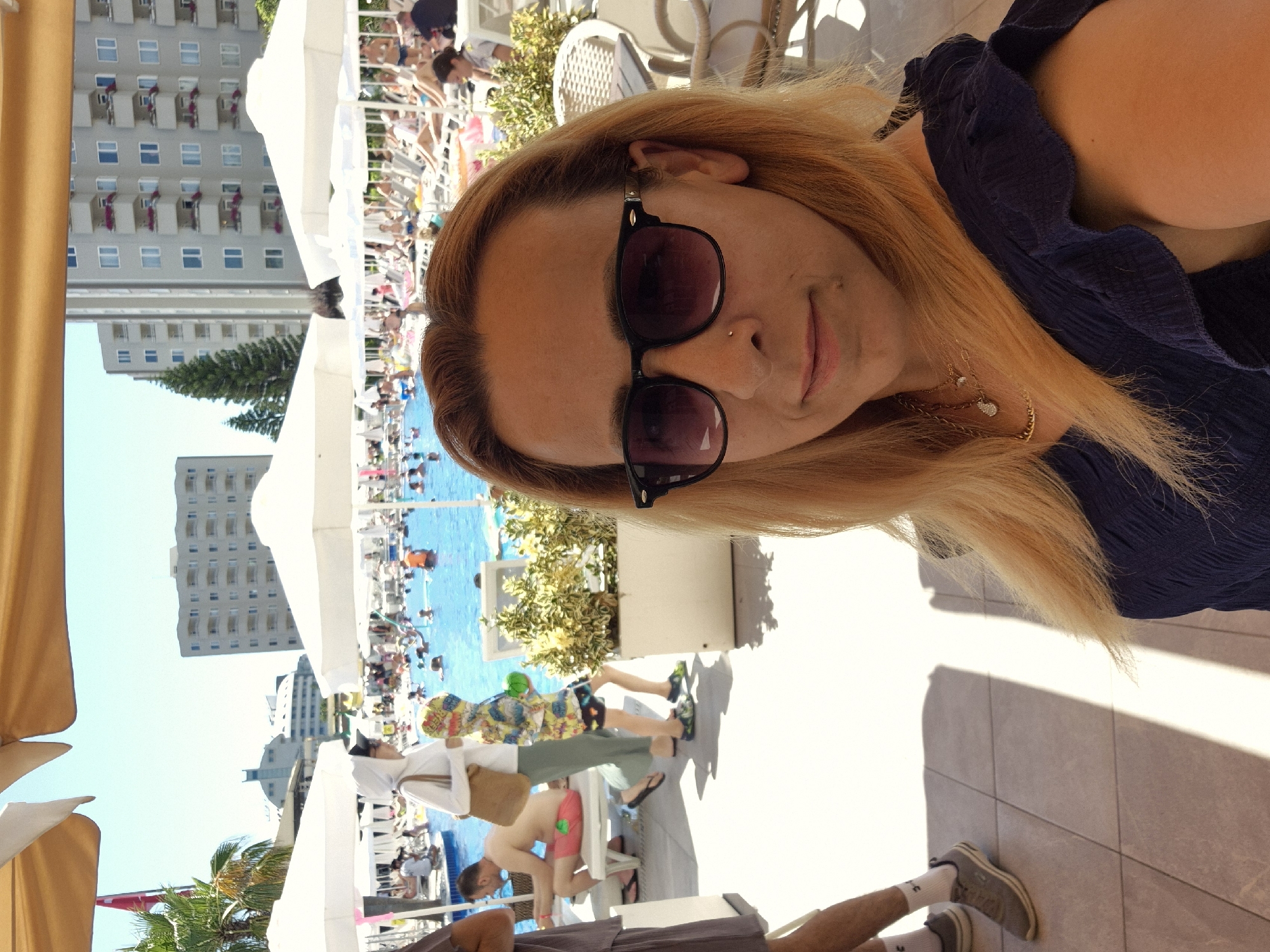 The image shows a woman with blonde hair and sunglasses taking a selfie in an outdoor setting. She is wearing a dark blue top and a necklace. The background features a swimming pool with people in it, lounge chairs, umbrellas, and buildings. Sunlight suggests it is daytime, and the scene appears to be a resort or hotel pool area. There are people in swimsuits and casual clothing, some relaxing, others in the pool. The overall atmosphere seems relaxed and summery. The image is oriented sideways, requiring rotation for proper viewing.