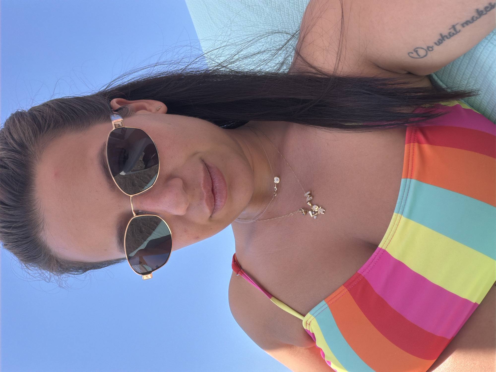 A woman is pictured outdoors under a clear blue sky, likely in Burgas, Bulgaria, as she reclines on a turquoise surface, possibly a beach chair. She is wearing a colorful, striped bikini top. Her right arm is visible, and it features a tattoo on her bicep that reads "Do what makes you happy." She is also wearing sunglasses and two delicate necklaces, one with a small diamond pendant and another with a charm resembling a cluster of figures. Her hair is dark and long, falling over her shoulder. The lighting suggests it is daytime, with the sun casting a slight shadow on her chest.