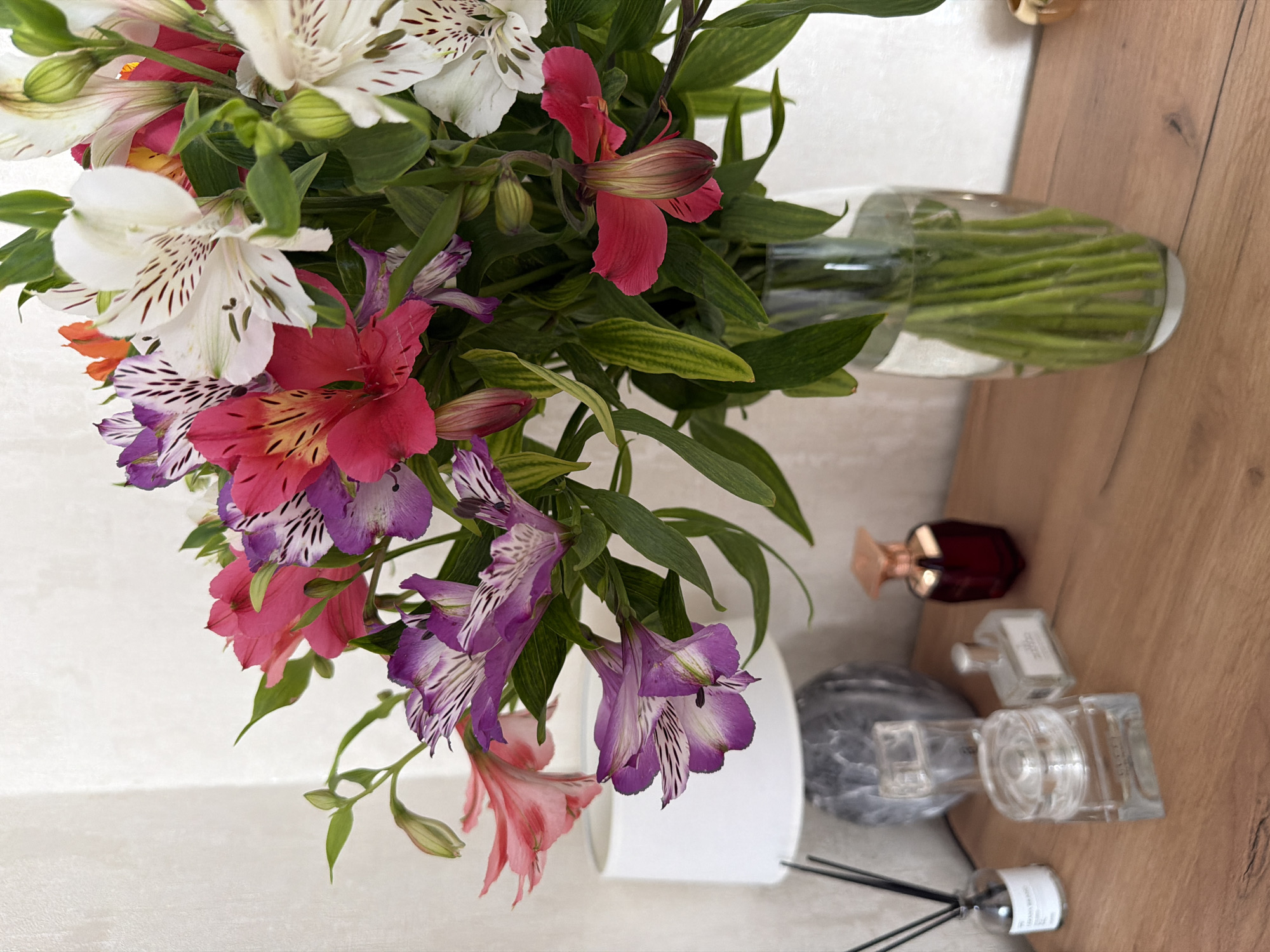 The image shows a vase of colorful alstroemeria flowers with white, pink, red, and purple blossoms. The flowers are in a clear glass vase with green stems visible. Adjacent to the vase, on a wooden surface, are various perfume bottles and a diffuser. The background consists of a plain white wall. The scene appears to be set indoors.