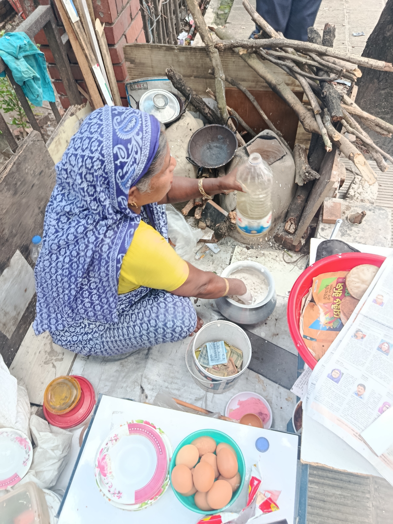 A woman in Dhaka, Bangladesh, wearing a blue patterned headscarf and yellow shirt, sits on the ground, preparing food. She mixes ingredients in a metal pot while pouring liquid from a clear plastic bottle. Next to her is a bucket filled with cash, suggesting she is selling the food. In front of her, on a white surface, are plates and a bowl of eggs. Behind her, firewood is stacked next to a cooking area with a pan on top. A red bowl filled with food and newspaper is also visible. The overall scene depicts a street food vendor at work.