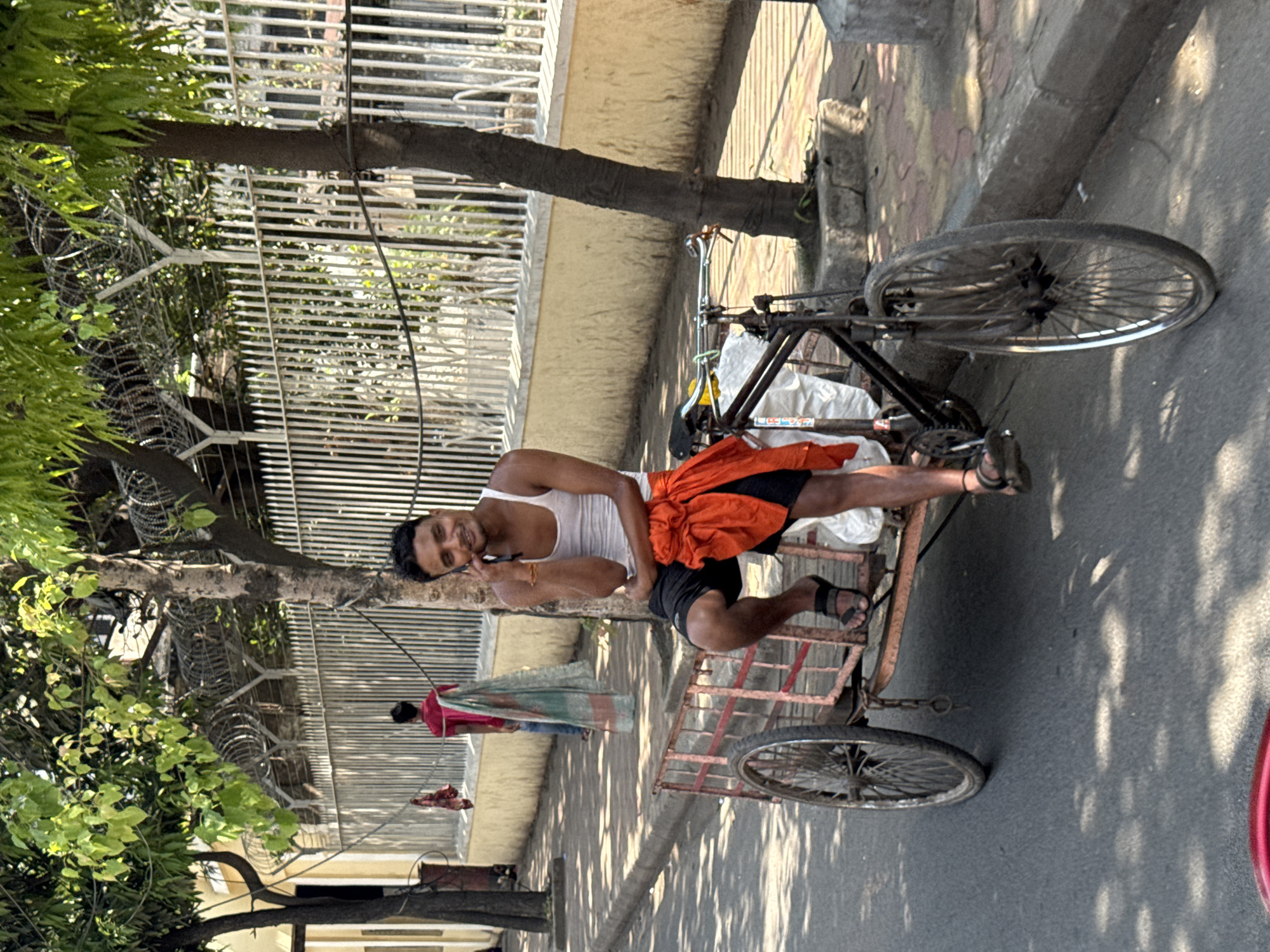 The image depicts a daytime street scene in Kolkata, India. A young adult male is seated on the forward edge of the cargo platform of a three-wheeled cycle rickshaw, positioned on an asphalt road near a concrete curb. He is wearing a white sleeveless top, black shorts, and dark sandals, with a bright orange fabric draped over his lap. The man holds a dark mobile phone to his right ear with his right hand, engaged in a call, and is smiling towards the viewer. The cycle rickshaw features a black bicycle front attached to a reddish-brown, open-sided metal cargo platform with two rear wheels.
Behind the main subject, a paved sidewalk runs parallel to the road, bordered by a low, cream-colored wall. Above this wall, a tall white metal fence with vertical bars extends upwards. Multiple strands of coiled razor wire are secured along the top of the fence, forming a security barrier. Several trees with green foliage line the street, casting dappled shadows on the road. Further down the sidewalk, another male figure is visible from behind, wearing a red shirt and blue pants, and carrying a light-colored fabric over his shoulder, walking away from the camera. A piece of red fabric hangs from a wire or line above the sidewalk in the mid-ground.