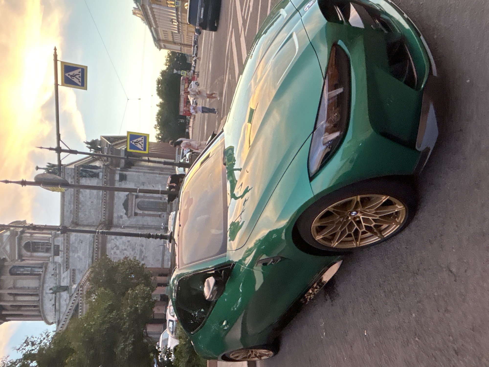 A bright green BMW M4 coupe is parked on a city street. The car is a coupe, with a low-slung profile and gold alloy wheels. The hood of the car is long and sculpted, with subtle creases. The headlights are narrow and angular, with LED daytime running lights. The car's bodywork is smooth and flowing, with a dramatic silhouette.
In the background, a large, ornate building with classical architecture can be seen. The building is made of stone, with tall columns and arched windows. The sky is a pale blue, with streaks of orange and yellow clouds, suggesting it might be sunset. Pedestrian crossing signs are visible on poles in the background. There are several people walking in the distance, as well as a red double-decker bus. The street is paved with asphalt, and there are some trees and other cars visible.