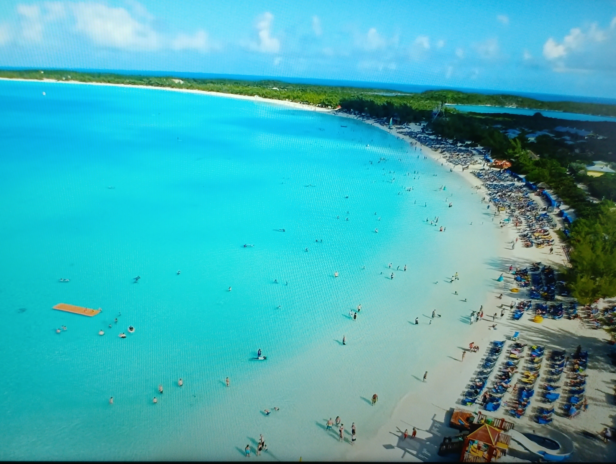 An aerial shot captures a vibrant beach scene under a bright, cloudy sky. The turquoise water is crystal clear, revealing the sandy seabed beneath. Numerous people are scattered across the water, some swimming, others wading, and a few lounging on an orange floating mat.
The white sandy beach is lined with lush green trees and packed with rows of blue lounge chairs, many occupied by sunbathers. A playground structure with slides is visible near the shore. The shoreline stretches into the distance, meeting a verdant landscape of trees and distant hills. The scene evokes a sense of tropical paradise and leisure.
This image is not related to the city of Dubăsari town council, Moldova.
