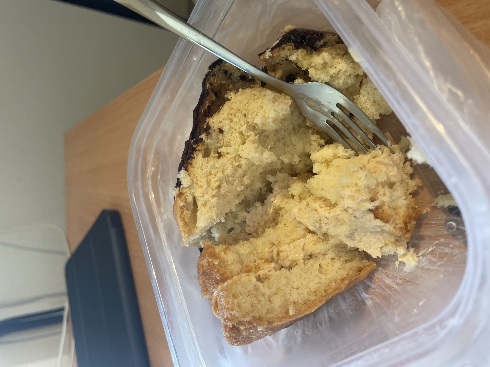 A close-up, top-down shot shows a slice of cake in a clear plastic container. The cake is yellow and appears crumbly, with some darker, possibly burnt, sections around the edges. A silver fork is inserted into the cake, with a few crumbs on the tines. The container is sitting on a light-colored wooden surface, with a blurred background that includes a dark electronic device and a lighter wall. The lighting suggests an indoor setting.