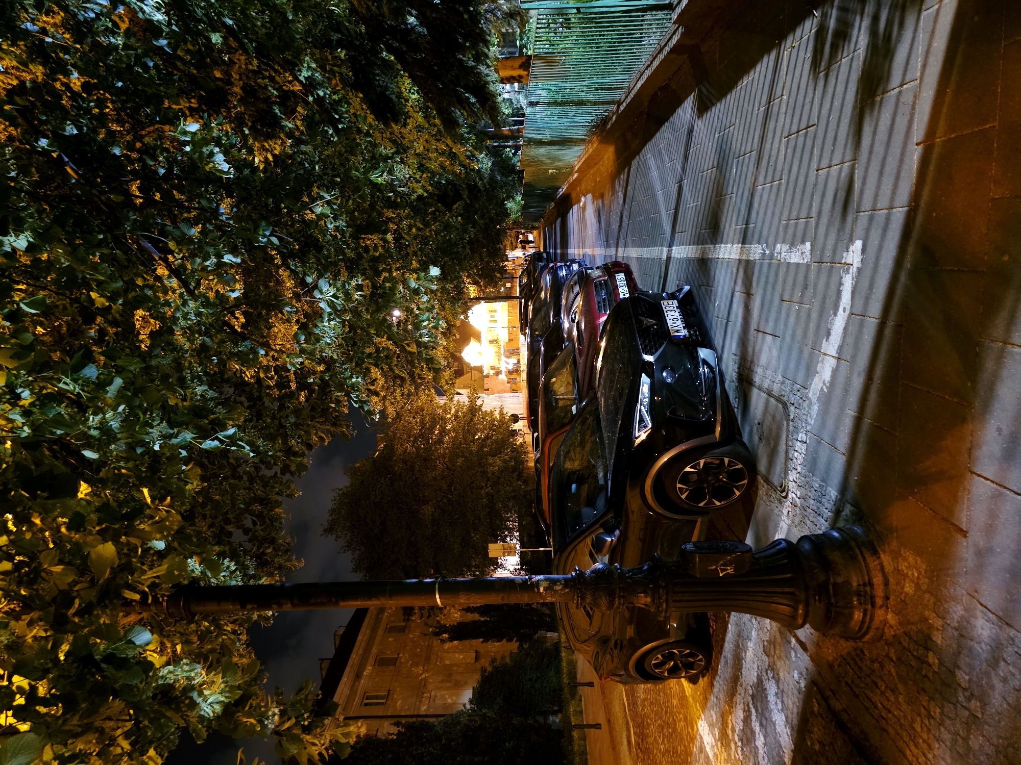 This image captures a street scene at night in Warsaw, Poland, likely in a residential or commercial area with trees lining the sidewalk. The primary focus is on a row of parked cars, predominantly black and dark-colored SUVs, occupying designated parking spots. The street is paved with cobblestones and marked with white lines. A prominent black lamppost stands in the foreground, casting shadows. Lush green trees with dense foliage arch over the scene, illuminated by warm-toned streetlights, creating a dappled effect on the ground and cars. In the background, a building with illuminated windows suggests habitation or businesses. A green metal fence runs along the right side of the image, bordering a grassy area or another part of the street. A few pedestrians are visible in the distance, walking along the sidewalk. The overall atmosphere is quiet and nocturnal, with the artificial lights interacting with the natural elements of the trees.