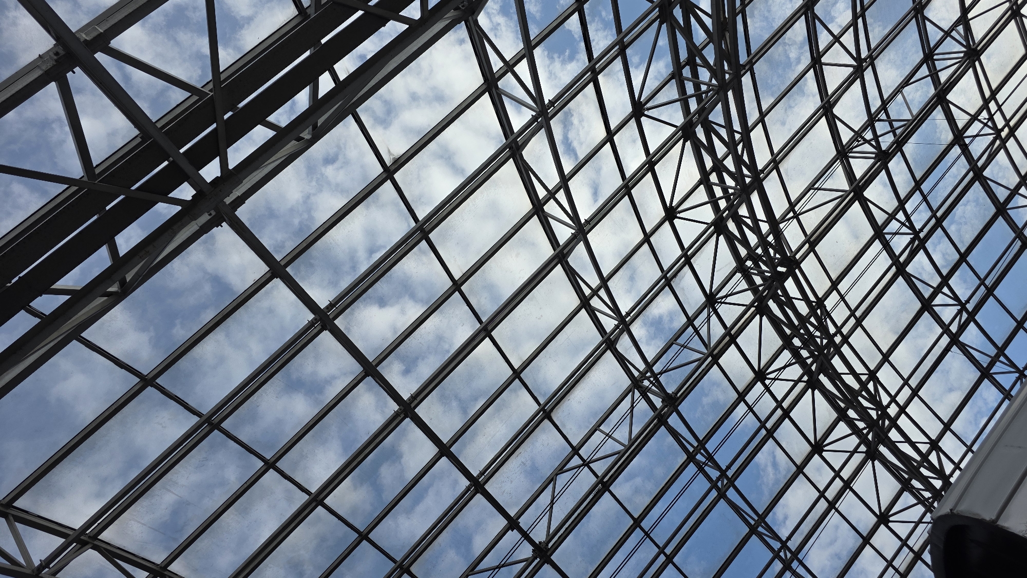 The image is a low-angle shot looking up at a large, industrial-style glass and steel roof structure. The roof is composed of a complex lattice of dark metal beams supporting translucent panels, through which a blue sky with scattered white clouds is visible. The perspective creates a sense of depth and a geometric pattern formed by the intersecting beams. The lighting suggests it is daytime, with natural light filtering through the roof. There are no people or specific identifiable landmarks that would indicate a location in Brest, Belarus. The scene is architectural and abstract, focusing on the structure of the roof and the sky beyond.