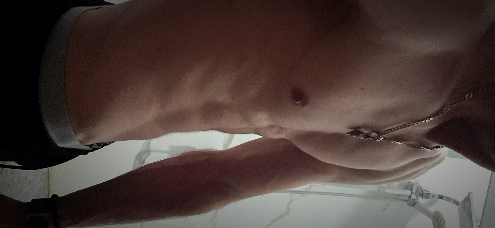 A torso, arm, and necklace are visible in this close-up, angled shot of a man. The individual has visible abdominal muscles. A gold chain necklace hangs on his chest. Part of a dark-colored garment and its grey waistband are seen on the left side of the frame. The setting seems to be a bathroom, indicated by the white tiled wall, showerhead, and faucet in the background. The exact location within the city of Plovdiv, Bulgaria, cannot be determined from this image alone.