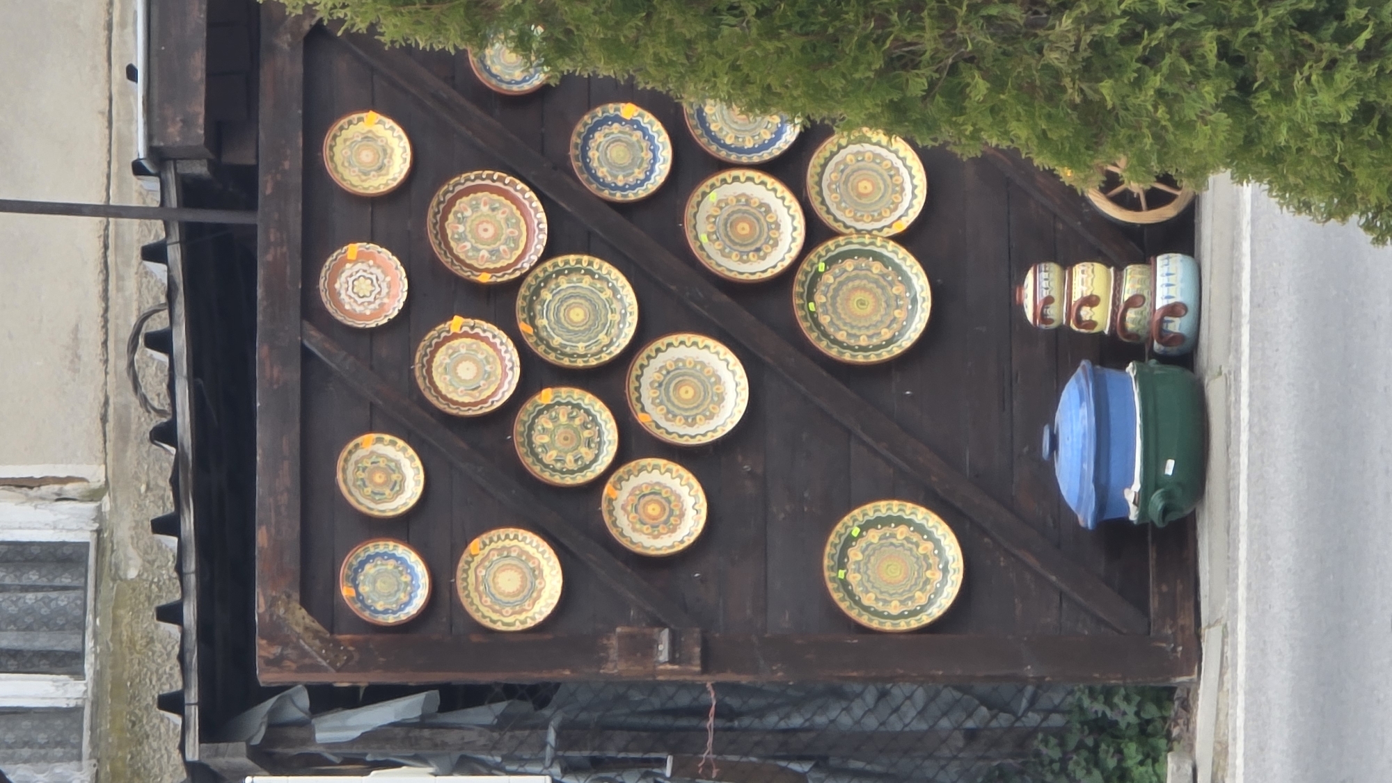 The image depicts an outdoor retail display of traditional Bulgarian pottery against a dark brown wooden gate or facade in the city of Rila, Bulgaria. Approximately 22 circular ceramic plates, ranging in size, are affixed to the wooden surface in a grid-like arrangement. Each plate features intricate, multi-colored geometric and floral patterns, predominantly in shades of green, yellow, brown, orange, and blue, with many displaying small, yellow circular price tags.
At the base of the wooden structure, on a narrow concrete ledge or sidewalk, a selection of larger ceramic vessels is displayed. To the left, a large, deep green ceramic pot with integrated handles is stacked beneath a matching blue ceramic lid, both showing a glossy finish. A white price label is attached to the green pot. To its right, a stack of three smaller, lidded ceramic bowls or pots, decreasing in size from bottom to top, exhibits varied glazes and hand-painted designs in light blue, yellow, and red-brown. Adjacent to this stack, a single light blue ceramic bowl with brown handles and a white design is visible. A segment of a wooden wagon wheel is partially visible behind these items on the right.
The wooden display structure itself consists of dark brown planks, some arranged diagonally, suggesting a gate or large double door. Metal corner brackets are visible at the top left. Above the wooden structure, an older, light-colored building is partially visible, featuring a window with white frames and opaque, patterned lace curtains. A corrugated dark metal roof section is situated between the window and the wooden display.
To the far left, a grey tarp or fabric is draped over an unseen object, and a section of a wire mesh fence is partially discernible. On the far right, a dense, bright green coniferous bush or tree occupies approximately one-third of the frame vertically, partially obscuring the pottery display and the wooden wheel. The foreground shows a grey asphalt road or street, separated from the pottery display by a concrete curb. No individuals are present in the scene.