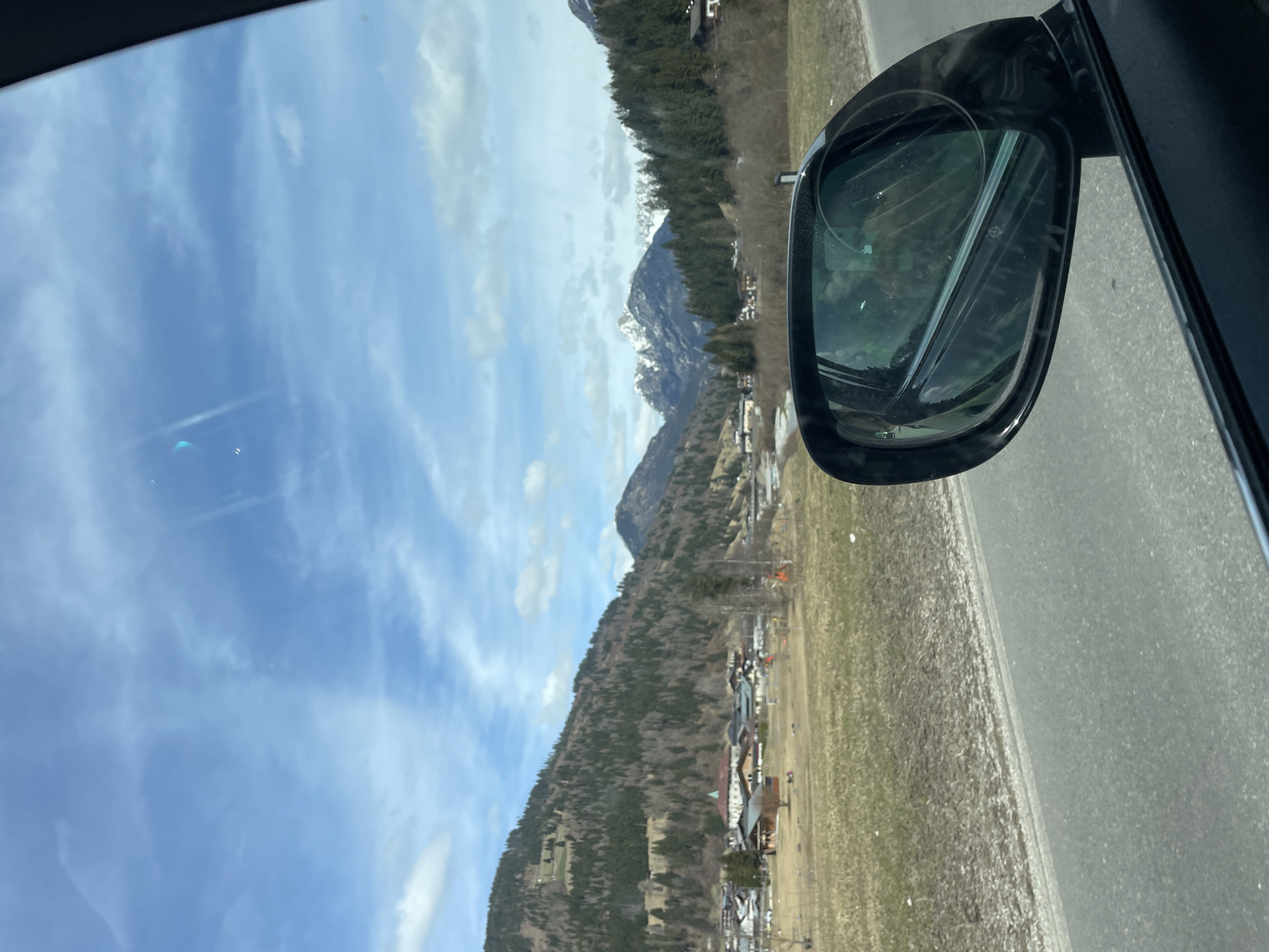 The image shows a view from inside a car, looking out towards a mountainous landscape. The car's side mirror is visible in the foreground, reflecting the scenery. Outside, there are mountains covered with trees, some snow-capped peaks, and a town nestled in the valley below. The sky is blue with scattered clouds. The scene is in Santo Stefano di Cadore, Italy.