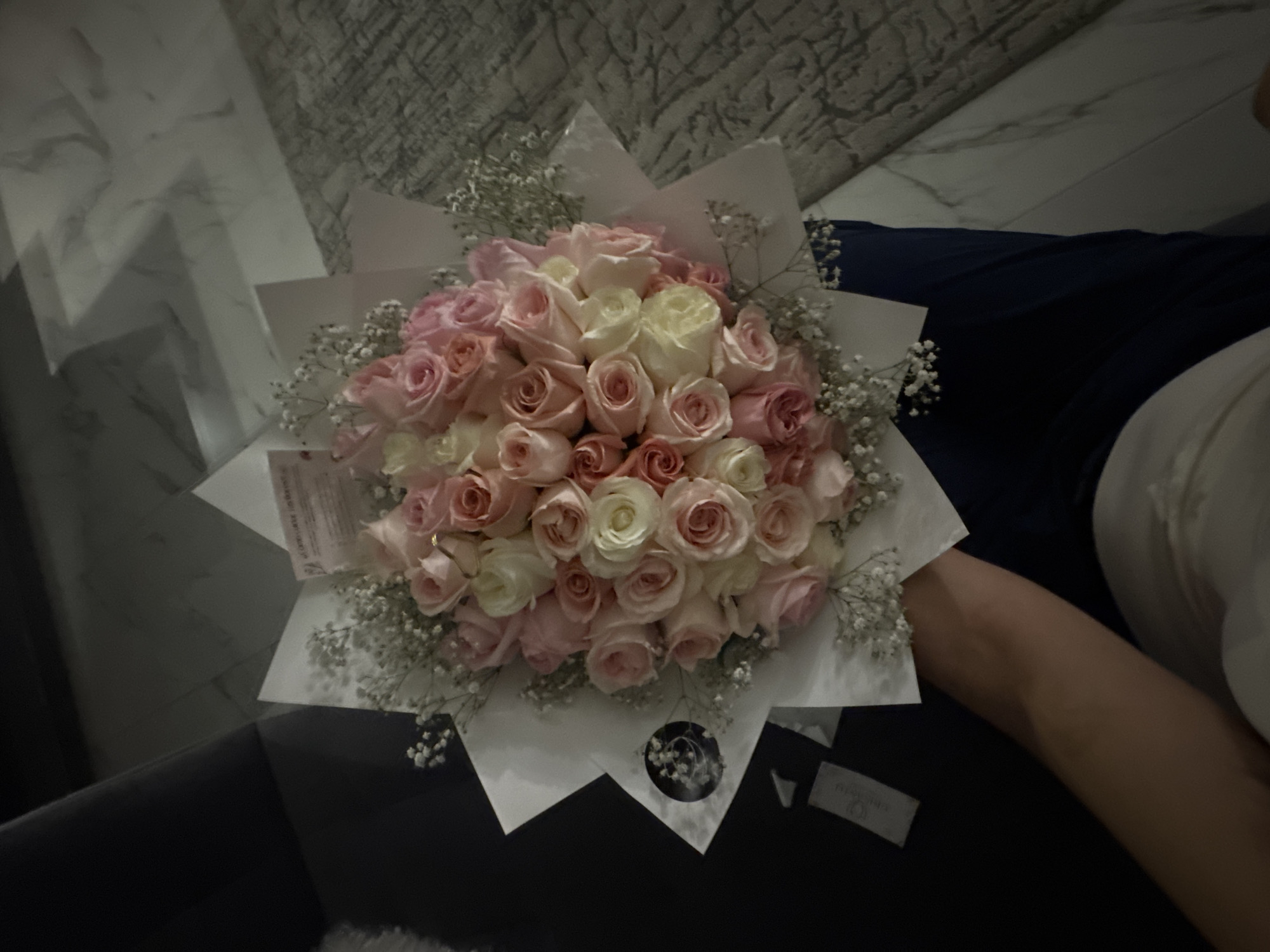 Here's a detailed description of the image:
**General Description:** The image features a bouquet of pink and white roses with baby's breath, wrapped in white paper. The bouquet is positioned on a dark surface, likely a sofa or chair. A person's arm is visible in the lower right portion of the image.
**Subjects:**
* **Bouquet:** The focal point, consisting of roses and baby's breath in a wrapping.
* **Person's Arm:** Partially visible, suggesting someone is holding or near the bouquet.
**Objects:**
* **Roses:** Pink and white roses that form the main part of the bouquet.
* **Baby's Breath:** Small white flowers used as filler in the bouquet.
* **Wrapping Paper:** White paper used to package the flowers.
* **Sofa/Chair:** The dark surface on which the bouquet rests.
**Environment:**
* **Interior:** The setting appears to be indoors, likely a home or apartment, based on the sofa/chair and the background (which shows hints of a marble-like surface and a textured material, perhaps a wall covering or another piece of furniture).
**Location:**
There is no concrete evidence to suggest that the photograph was taken in Puebla, Mexico.