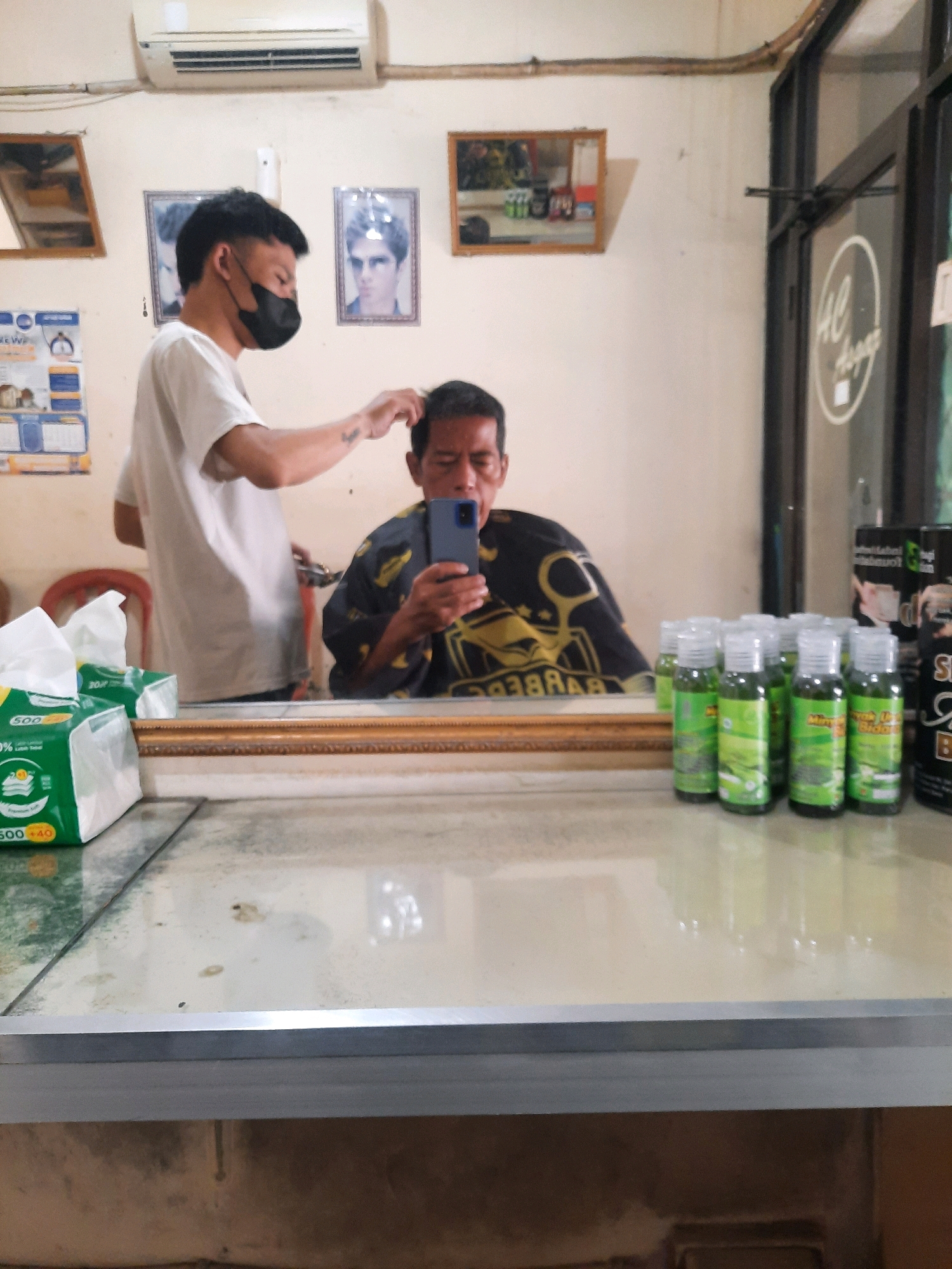 In East Jakarta, Indonesia, a barber is cutting the hair of a man in a barbershop. The barber, wearing a white t-shirt and a black mask, is using clippers on the man's head. The man, seated and wearing a patterned cape, is looking at his phone. On the counter in front of them are bottles of hair product and a box of tissues. In the background, framed photos are hanging on the wall, and an air conditioner is mounted above. The scene is reflected in a large mirror behind the counter.