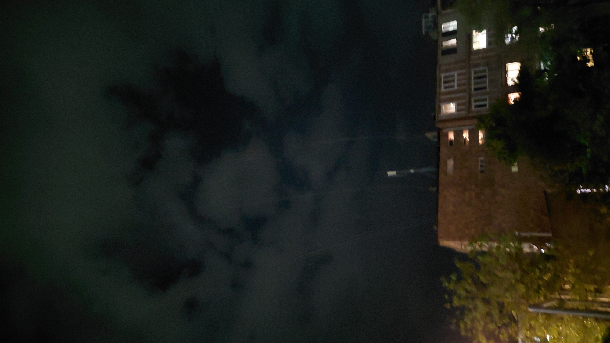 The image depicts a nighttime scene in the city of Abovyan, Armenia, focusing on an apartment building. The sky is dark with clouds, some of which are illuminated by faint light. Several thin, light-colored lines, possibly wires or thin beams of light, extend downwards from the sky. The building in the foreground is multi-story with a brick facade on the left side and lighter colored sections on the right. Numerous windows are visible, some of which are lit from within, indicating occupancy. Trees with green foliage partially obscure the lower portions of the building, suggesting the scene is viewed from a street or park area. The overall impression is one of an urban residential environment at night.