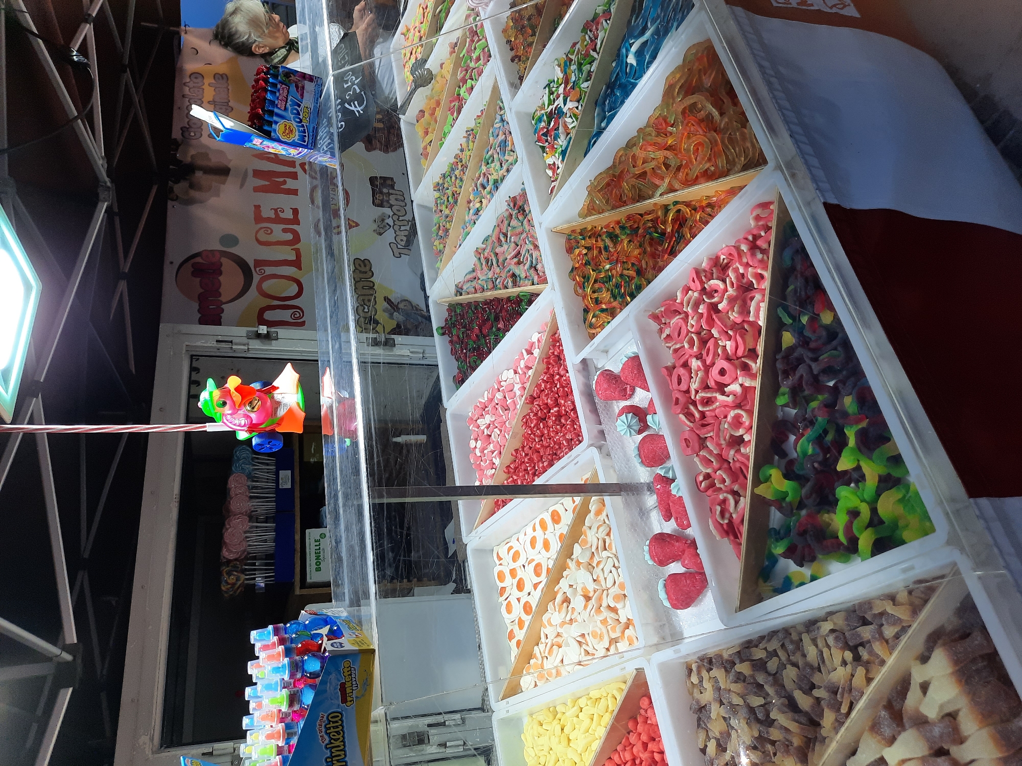 An outdoor candy stand in Pesaro, Italy, displays a variety of colorful candies. The stand is covered by a white canopy. In the background, a woman with gray hair wearing glasses and a dark scarf is visible. She is leaning over the counter, her face obscured. A sign above the counter reads "DOLCE MA". The foreground is dominated by several clear plastic bins filled with different types of candy.
In the leftmost bin, yellow banana-shaped gummies are visible. To their right, a bin contains orange and white fried egg-shaped gummies. Next to that, a bin is filled with red heart-shaped candies. Further to the right, a bin holds an assortment of colorful gummy worms. Below these, bins contain brown and white cola bottle-shaped gummies, green and black gummy snakes, pink ring-shaped candies, and red strawberry-shaped gummies. There is also a bin of assorted candies in the upper right section of the image.
On the left side of the stand, a box of "Trinketto" flavored candy bottles is displayed. Behind the candy bins, rows of lollipops are arranged in blue containers. A colorful plastic toy character, with a green hat and pink and orange body, is suspended from a string above the stand. The overall scene depicts a vibrant and abundant candy market stall.