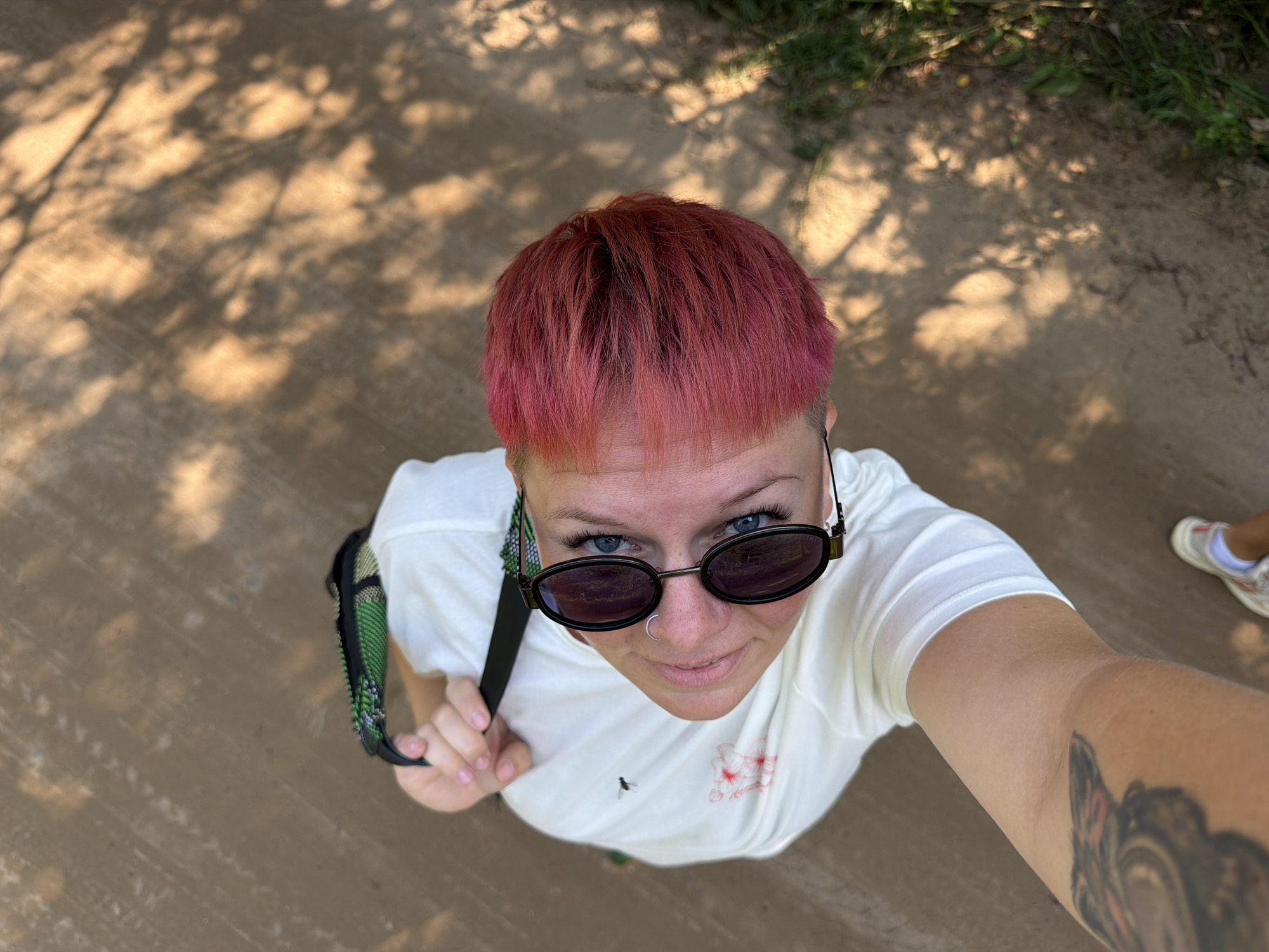 A high-angle, full-body shot captured from above shows a person with short, bright pink hair wearing a white t-shirt, black sunglasses, and a nose ring. They are holding a black strap with a green and black patterned bag over their shoulder. The person is standing on a dirt path with sunlight casting dappled shadows from trees. To the left of the frame, a tattoo is visible on their arm, and a pair of white sneakers with blue and white accents are visible on their feet. The scene appears to be outdoors, possibly in a park or a natural setting within the city of Chaikovsky, Russia, given the lighting and the presence of foliage.