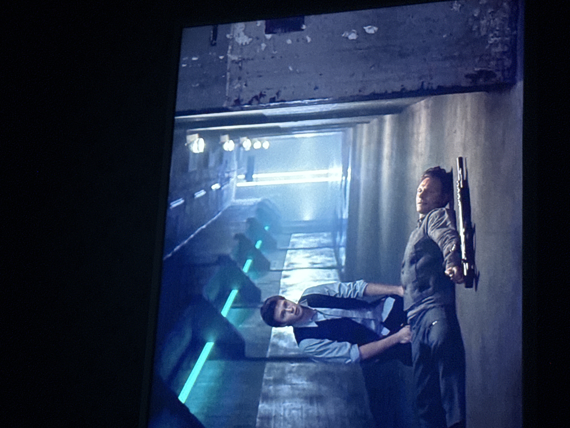 The image depicts a scene within a concrete tunnel. A man in a blue shirt and vest is shown lying on the ground, seemingly pushing the leg of another man. The second man is standing and leaning against the tunnel wall, holding what appears to be a rifle. The tunnel has a series of light fixtures along its length, creating a blue-lit environment. The location in the city of Mytishchi, Russia is not identifiable from the context of the image.