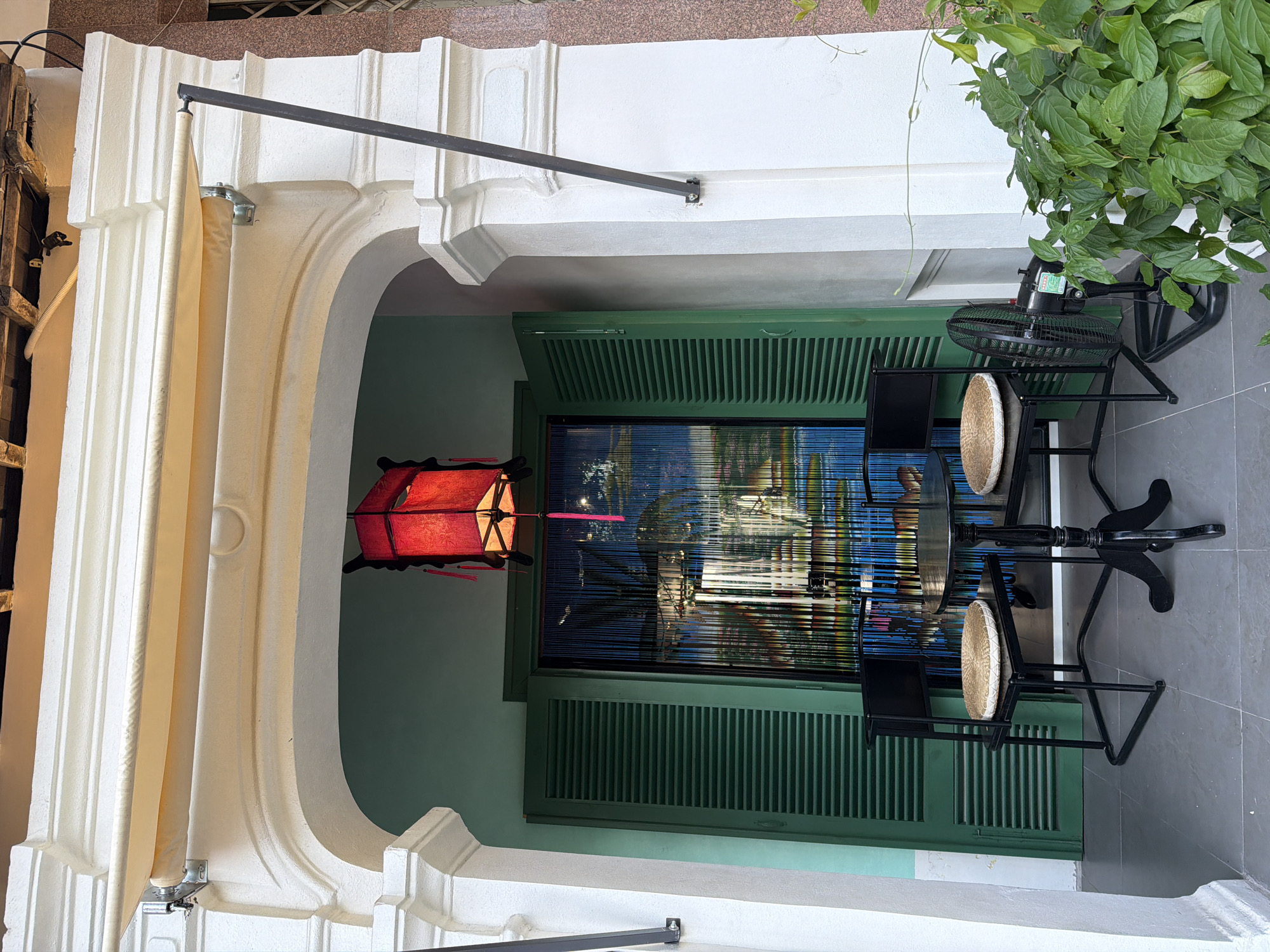 The image shows an outdoor seating area in front of a building. The area is covered by a white awning and features a round black table with two black chairs, each with a straw cushion. Behind the seating, there is a green louvered door and a doorway adorned with a colorful beaded curtain. A red lantern hangs from the ceiling above the doorway. To the right of the seating area, a black electric fan stands on the ground next to lush green foliage. The building's facade is white with ornate architectural details. The scene is likely a cafe or restaurant entrance in Nha Trang, Vietnam, given the architectural style and the presence of outdoor seating. There are no people visible in the image.