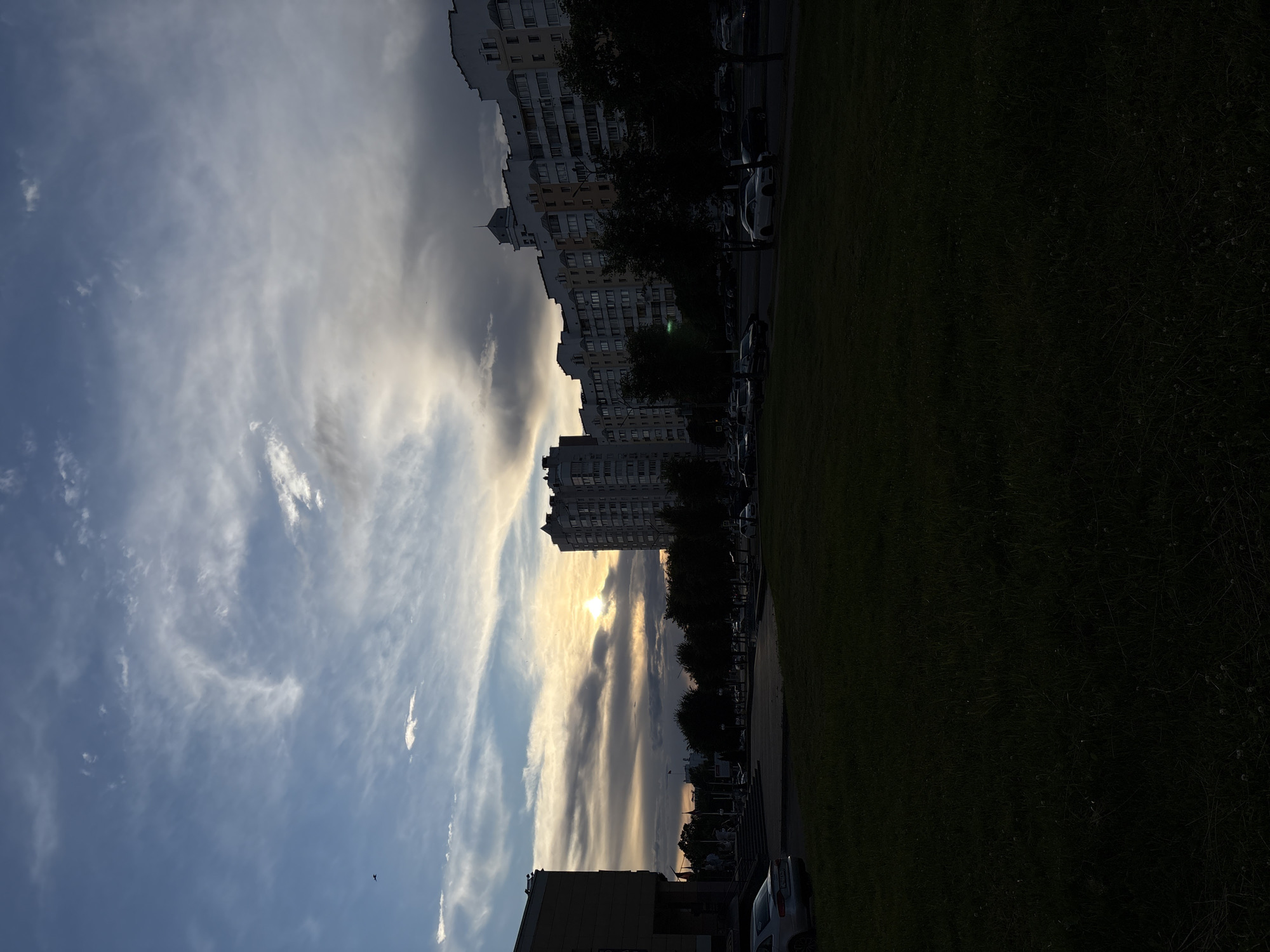 This image depicts a street scene in Vitsebsk, Belarus, during sunset. The sky is filled with a mix of dramatic clouds, ranging from dark grey to lighter, wispy formations, illuminated by the setting sun which casts a warm glow. In the foreground, a dark, grassy area is visible, suggesting a park or a boulevard.
Dominating the background are multi-story residential buildings, characteristic of Soviet-era architecture, with their distinctive layered roofs and numerous windows. Trees line the street in front of the buildings, their foliage silhouetted against the sky. Several cars are parked along the roadside and appear to be in motion on the street, indicating normal urban activity.
On the left side of the image, part of a building with a light-colored facade is visible, featuring steps leading up to an entrance. A silver car is parked in front of this building. The overall impression is a city transitioning from day to night, with the natural beauty of the sunset contrasting with the urban landscape.