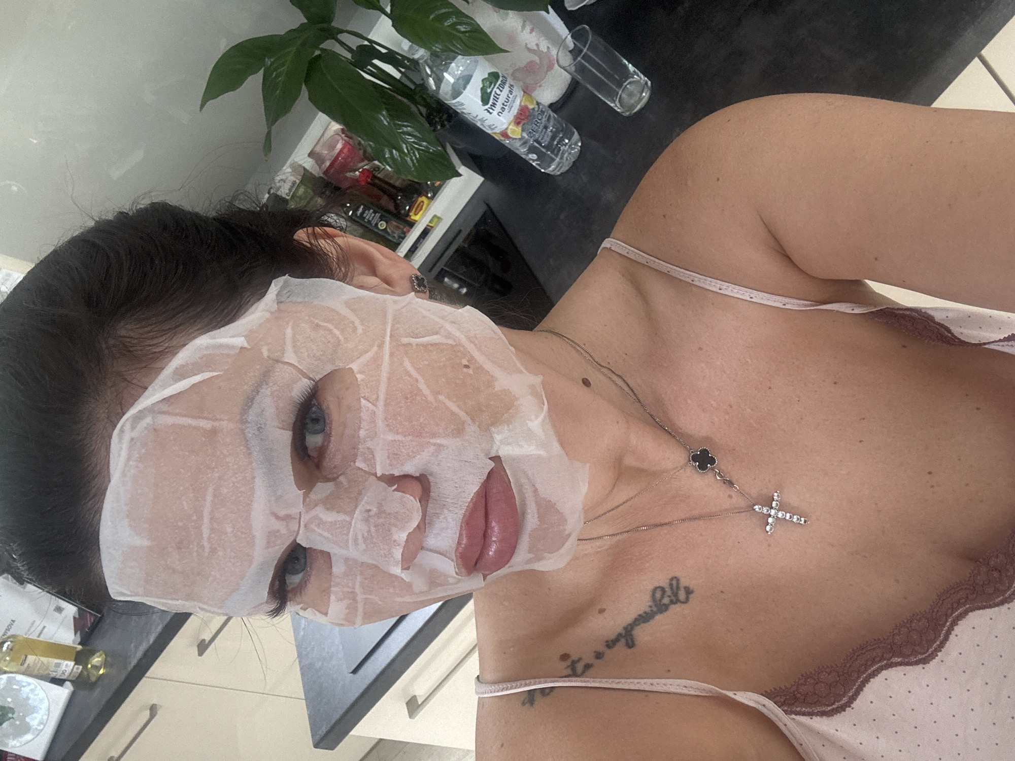 A woman with dark hair and a sheet mask on her face is taking a selfie in a kitchen. She is wearing a pink camisole with white polka dots and lace trim. A silver necklace with a cross pendant and a clover charm is visible on her chest. A tattoo is visible on her left shoulder and upper chest. In the background, there are kitchen cabinets, a countertop with bottles of liquids and a plant, and a glass. The scene is located in Katowice, Poland.