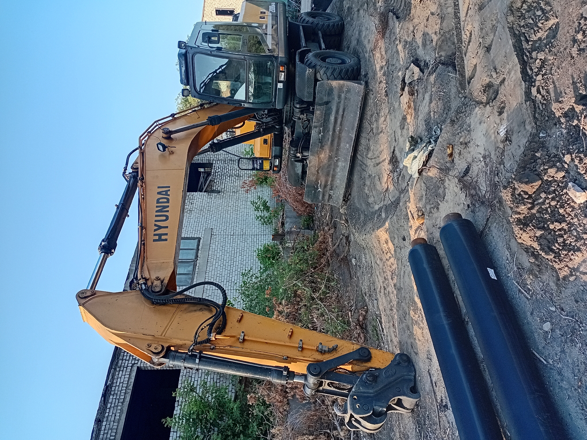 This image depicts an outdoor scene in the city of Semey, Kazakhstan, featuring a Hyundai wheeled excavator parked on a dirt construction site. The excavator is yellow and black, with a large bucket attachment. In the foreground, two black pipes are laid parallel on the ground. The background shows a weathered brick building with broken windows, overgrown vegetation, and debris scattered around the site. The sky is clear and blue, indicating daytime. There are no people visible in the image. The scene suggests a construction or demolition project is underway or has recently occurred.