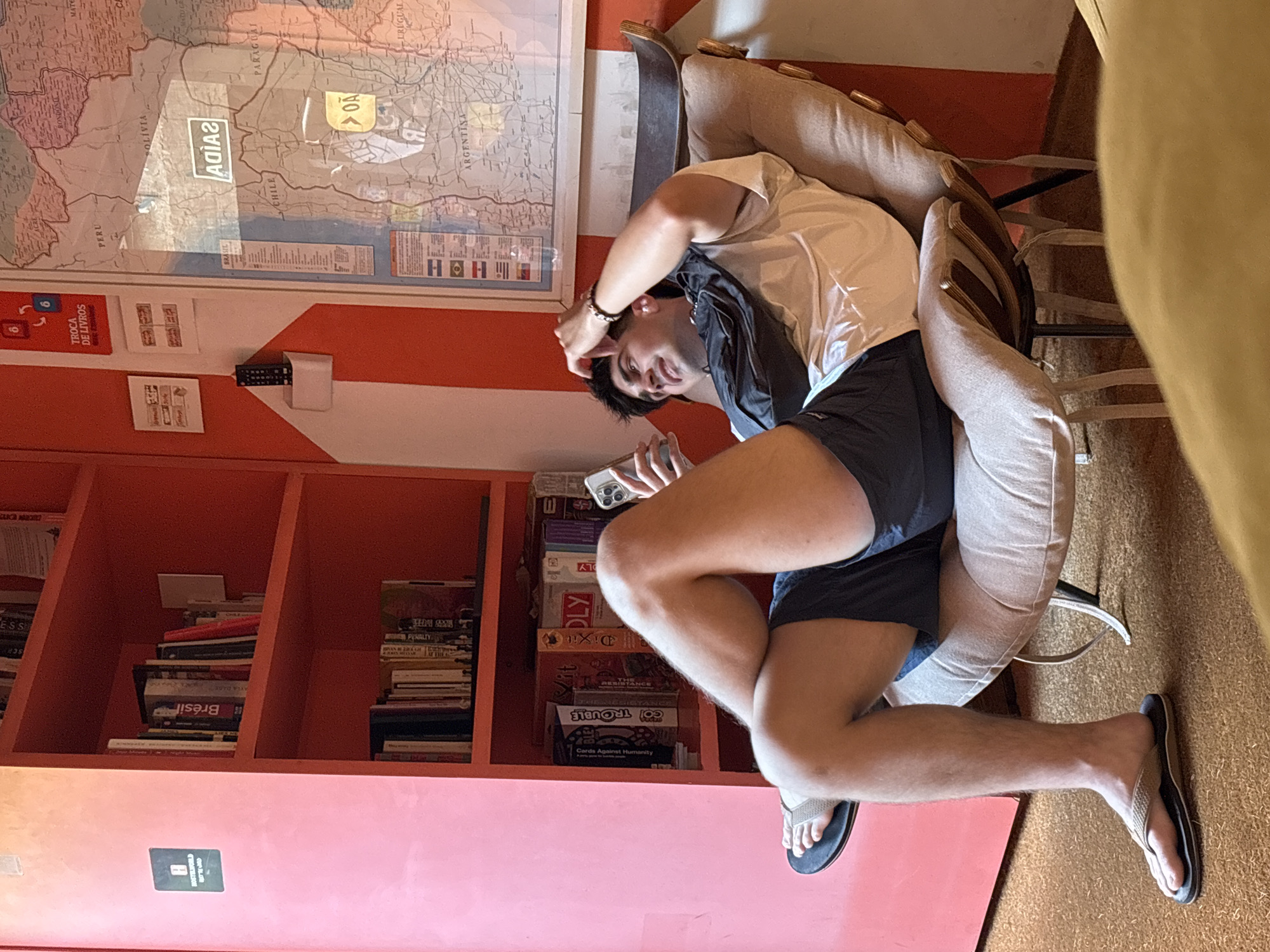 The image captures an indoor scene, likely a common area or lounge within a hostel, identified by a "TROCA DE LIVROS" (Book Exchange) sign and a prominent map. A young man, appearing relaxed, is the central subject. He is seated on a light-colored armchair, leaning back with his right arm raised towards his head, looking down at a smartphone held in his left hand. He wears a white t-shirt, dark shorts, and light-colored flip-flops.
The setting features warm orange walls, one of which displays a large map of South America. Visible countries on the map include BRASIL, PERU, CHILE, BOLIVIA, PARAGUAI, ARGENTINA, and URUGUAI. A reflected word "AGIAS" is visible on the map, which is likely "SAÍDA" (Portuguese for "exit") reflected from a sign outside the frame. Below the map, a remote control rests on a small shelf. Another orange wall features a bookshelf filled with books and various board games, such as "Dixit", "GO! TROUBLE", "THE RESISTANCE", and "Cards Against Humanity". Some book spines are visible, including "Brésil" and "PENALTY". A small sign for "HOSTELWORLD" is also visible on the lower shelf.
The lighting suggests it is daytime. The man's relaxed posture and the presence of amenities like a book exchange and board games contribute to an atmosphere of leisure within what appears to be a communal space in São Paulo, Brazil.
