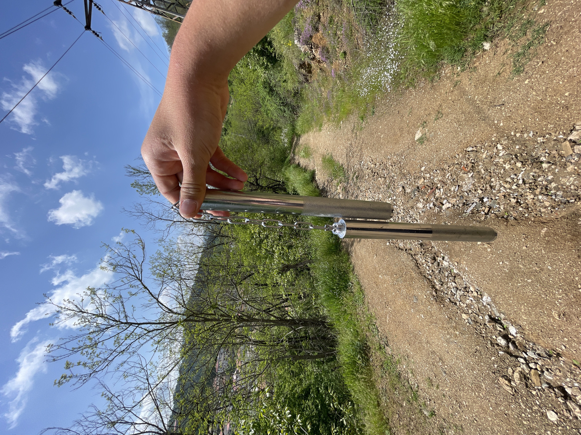 A hand holds metal tubes attached by a chain and circular ornament against a natural background. The background is a dirt trail lined with green vegetation, trees, and a distant hillside. The sky is blue with scattered clouds. The location is Zlatograd, Bulgaria.