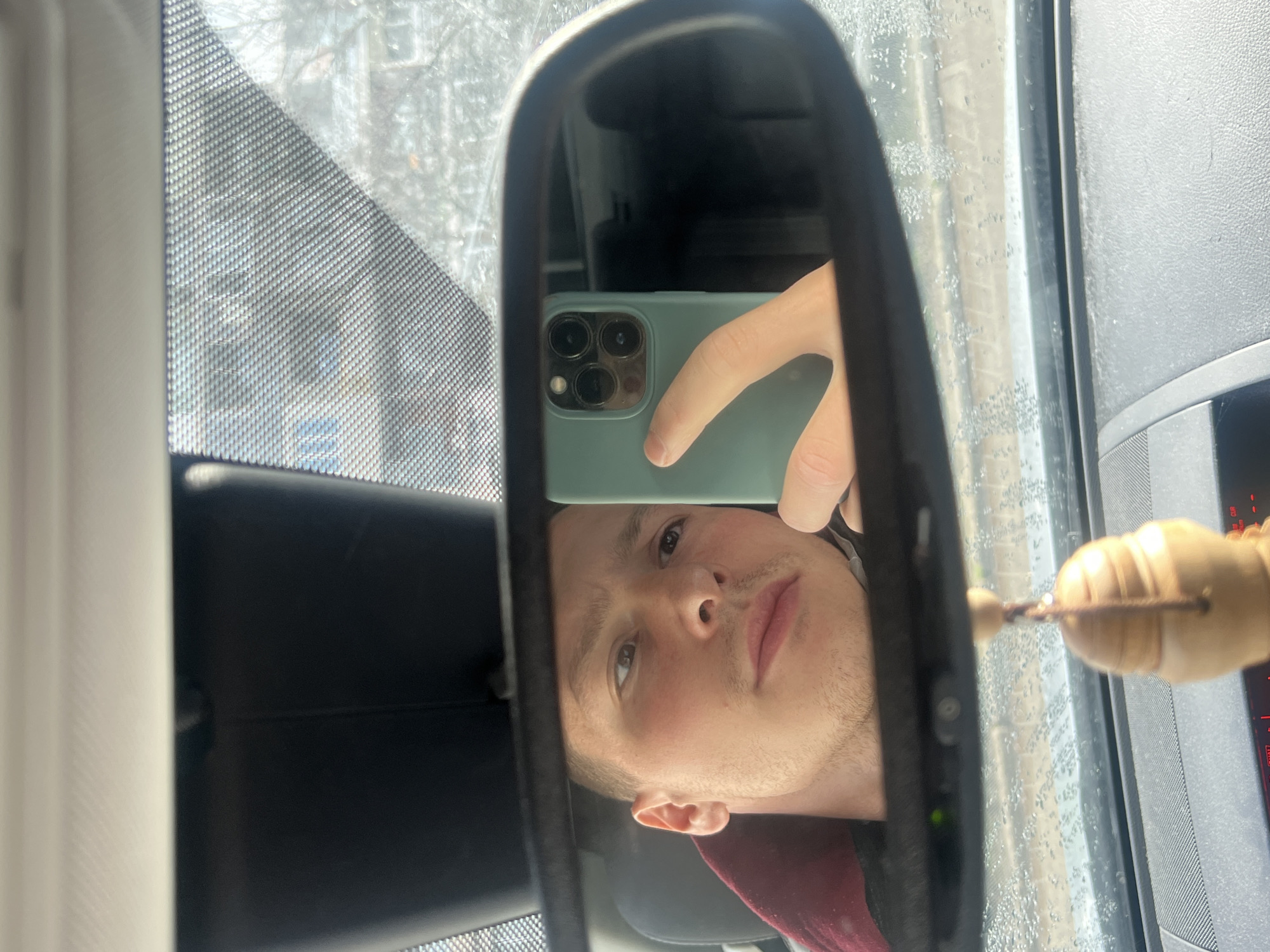 A person is taking a selfie in the rearview mirror of a car. The phone is held up to the mirror, partially obscuring the person's face. The person has light skin, short hair, and is wearing a red shirt. A string of wooden beads hangs from the mirror. Outside the car window, there are trees, buildings, and other cars, suggesting a street scene. The specific location in the city of Burgas, Bulgaria cannot be determined from the image.