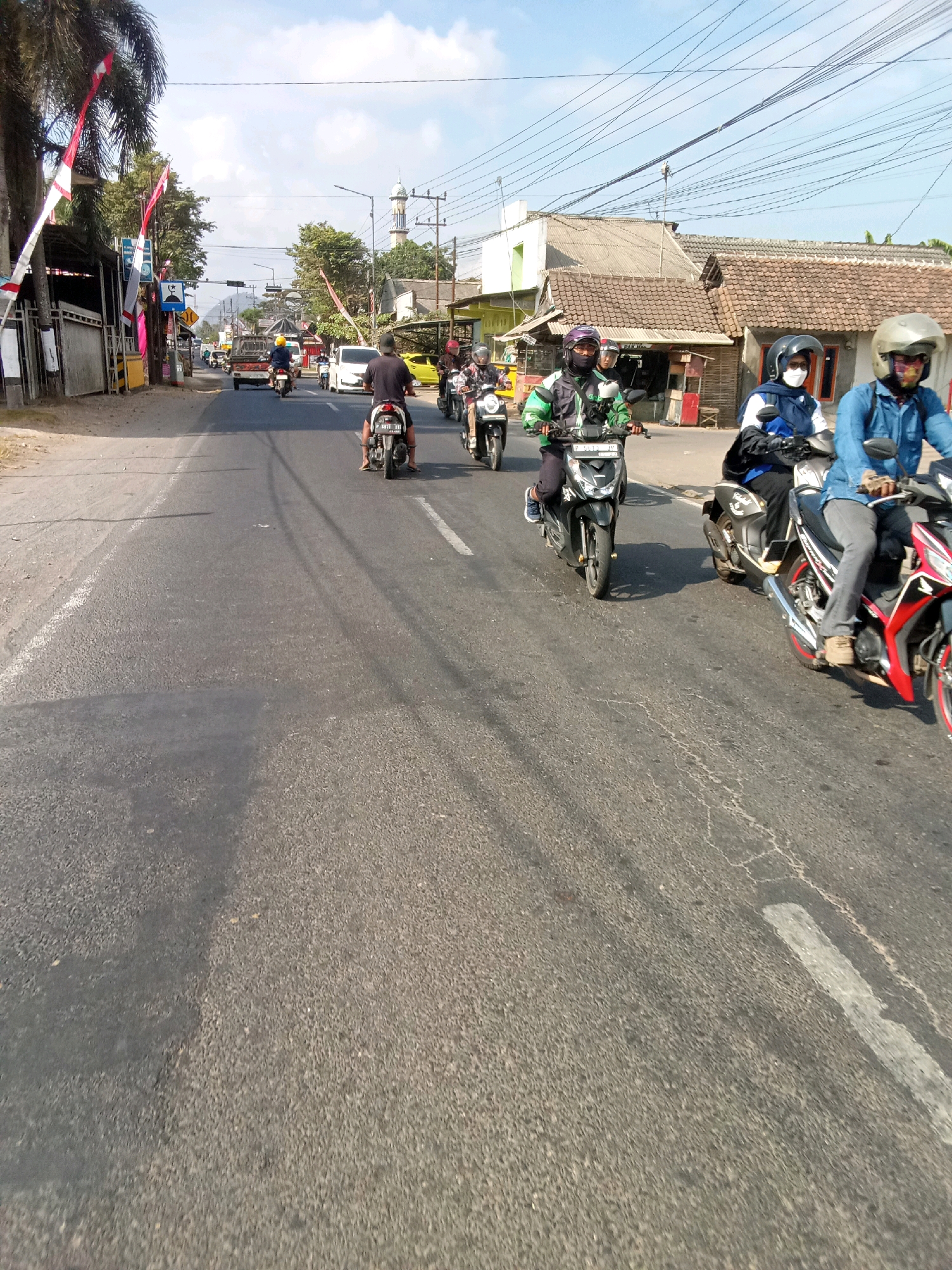 The image depicts a street scene in Klompangan, Indonesia, on a sunny day. Several motorcycles are navigating the asphalt road, with riders wearing helmets for safety. Some motorcycles are traveling in a group, suggesting a convoy or a shared journey. The road is lined with buildings, including houses and shops, with trees and vegetation visible in the background. Indonesian flags and banners adorn some of the buildings, indicating a possible local celebration or event. Power lines crisscross the sky, typical of an urban or semi-urban environment. The overall atmosphere is one of daily life and movement on the road.