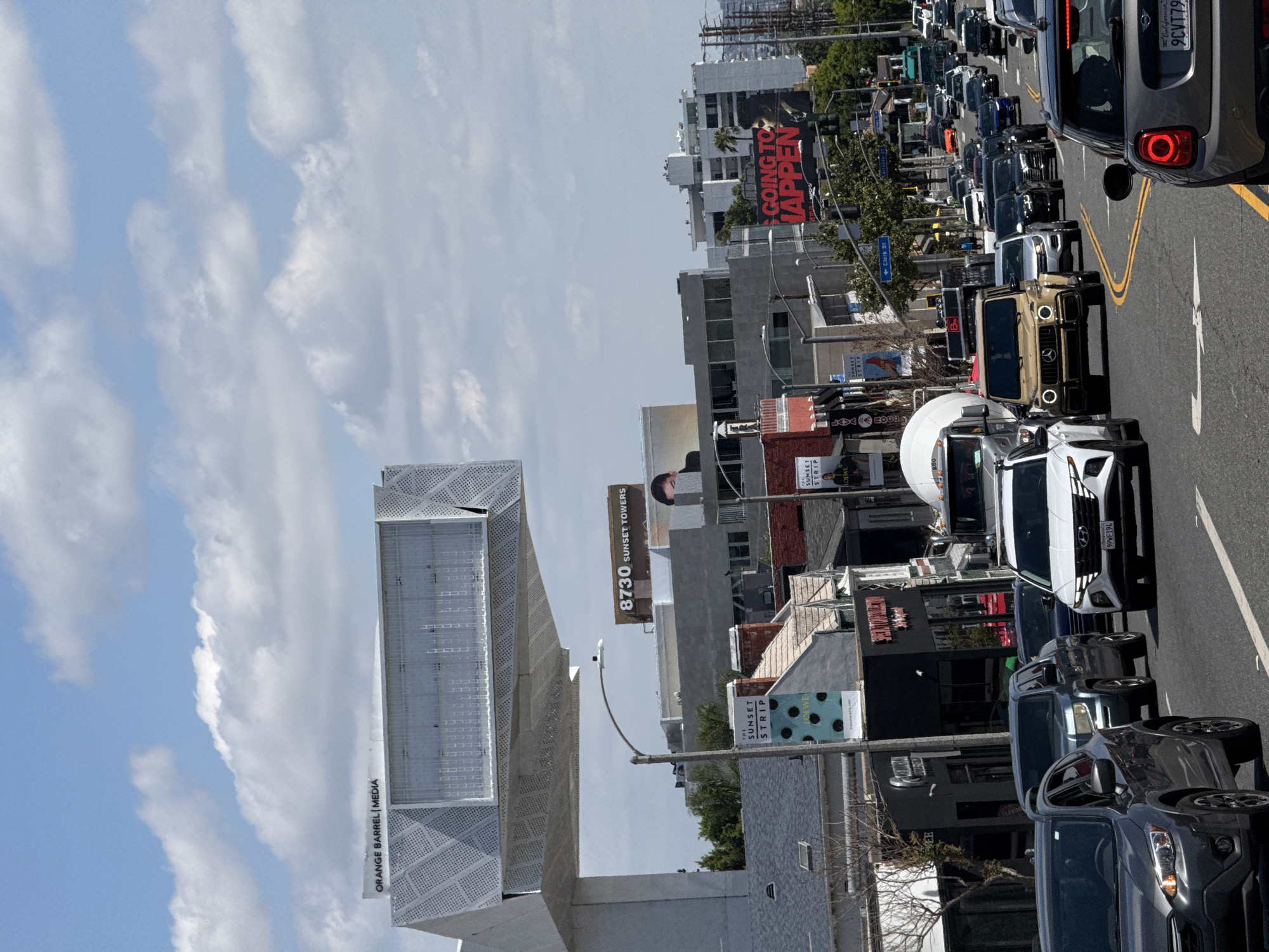 This image is a street scene in West Hollywood, United States of America, showing a busy street with heavy traffic. Prominent landmarks include the ORANGE BARREL | MEDIA structure, a billboard advertising 8730 SUNSET TOWERS, and a large advertisement that reads "GOING TO HAPPEN". Various businesses are visible along the street, indicated by signs like "THE SUNSET STRIP" and "LOEWE". The street is densely packed with cars, primarily facing the same direction, with visible license plates on some vehicles. The sky is partly cloudy, and there are poles, wires, and trees alongside the road.