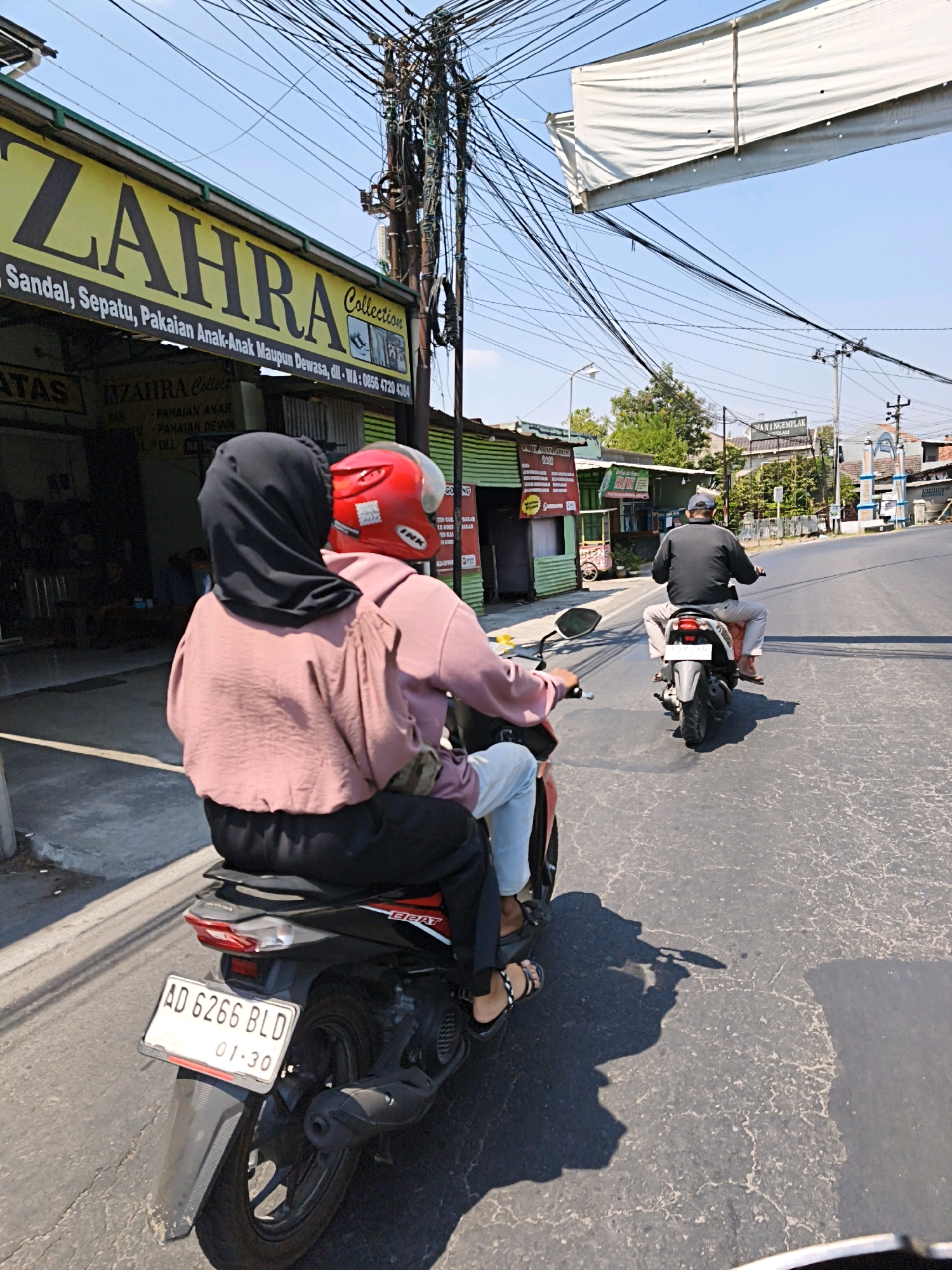 The image depicts a street scene in Desa Donohudan, Indonesia, on a sunny day with a clear blue sky. Two motorcycles are traveling on a paved road. In the foreground, a motorcycle carries two people. The rear passenger wears a black hijab and a pink long-sleeved shirt, while the rider wears a red helmet and a similar pink shirt. The motorcycle's license plate reads "AD 6266 BLD 01-30." The motorcycle in the background is ridden by a person wearing a dark long-sleeved shirt and light-colored pants, also with a helmet.
To the left of the road, a building with a yellow sign displaying "ZAHRA Collection" is visible. The sign also includes text in Indonesian indicating "Sandal, Sepatu, Pakaian Anak-Anak Maupun Dewasa, dll - WA : 0856 4720 4384." Below this sign, another smaller sign or banner also mentions "ZAHRA Collection" and lists items such as "Pakaian Anak" and "Pakaian Dewasa." Other shops or buildings with signs are present further down the street. The environment is characterized by a dense network of electrical wires overhead, characteristic of many urban and semi-urban areas in Indonesia. The road surface shows some wear and tear.
