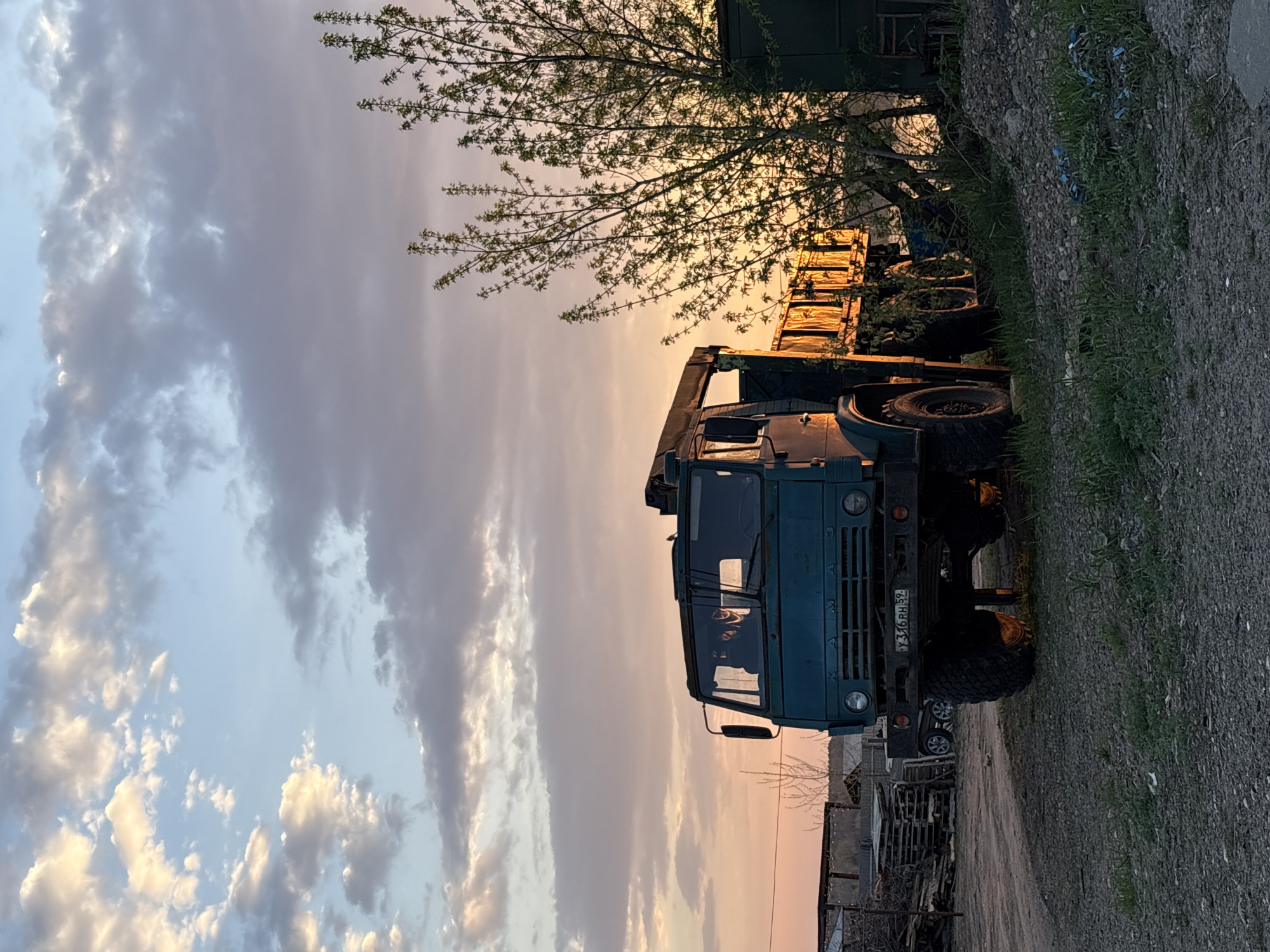 The image shows a blue KAMAZ truck parked near a road. The truck is in the city of Chaikovsky, Russia. There is a tree with budding leaves near the truck. The sky is filled with clouds illuminated by the setting sun. In the background, there is a building. Piles of wood are next to the road.