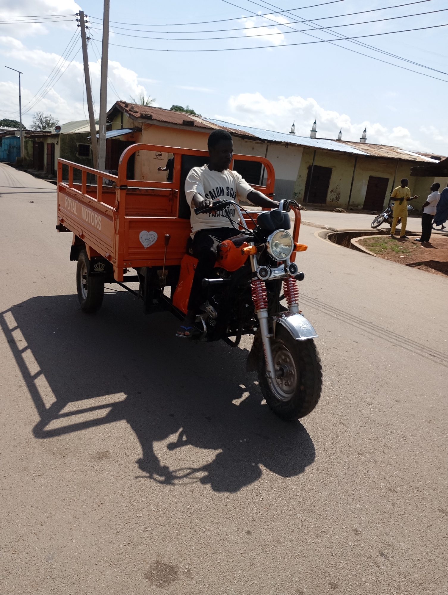 A man is riding an orange motorized tricycle down a paved road in Shendam, Nigeria. The tricycle has a flatbed cargo area at the back and a motorcycle front. The man is wearing a white t-shirt with text on it and dark pants. In the background, there are buildings with corrugated metal roofs, some of which appear to be shops. Power lines are visible overhead, supported by utility poles. Several other people are standing on the side of the road, and another motorcycle is parked nearby. The sky is partly cloudy. The scene suggests a typical day in a Nigerian urban or semi-urban setting.