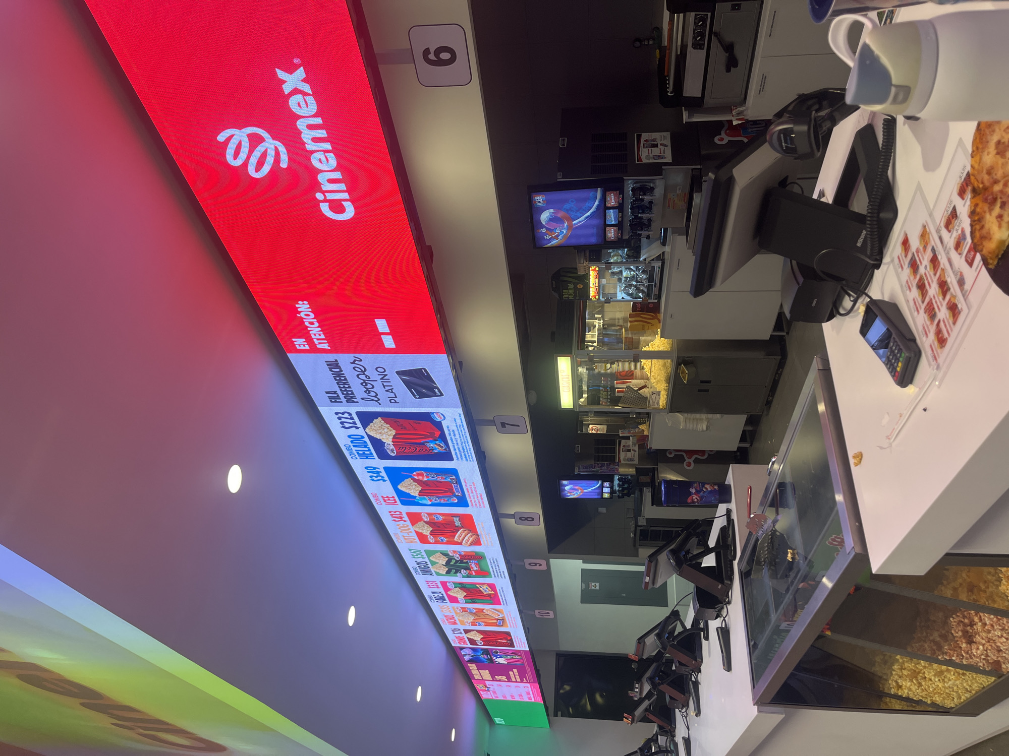 The image shows the interior of a Cinemex movie theater concession stand, likely in Heroica Puebla de Zaragoza, Mexico. The scene includes a red digital menu board displaying combo prices in pesos. Numbers mark the service points. POS systems are present at the counter. Popcorn and other snacks are visible behind a glass display case.