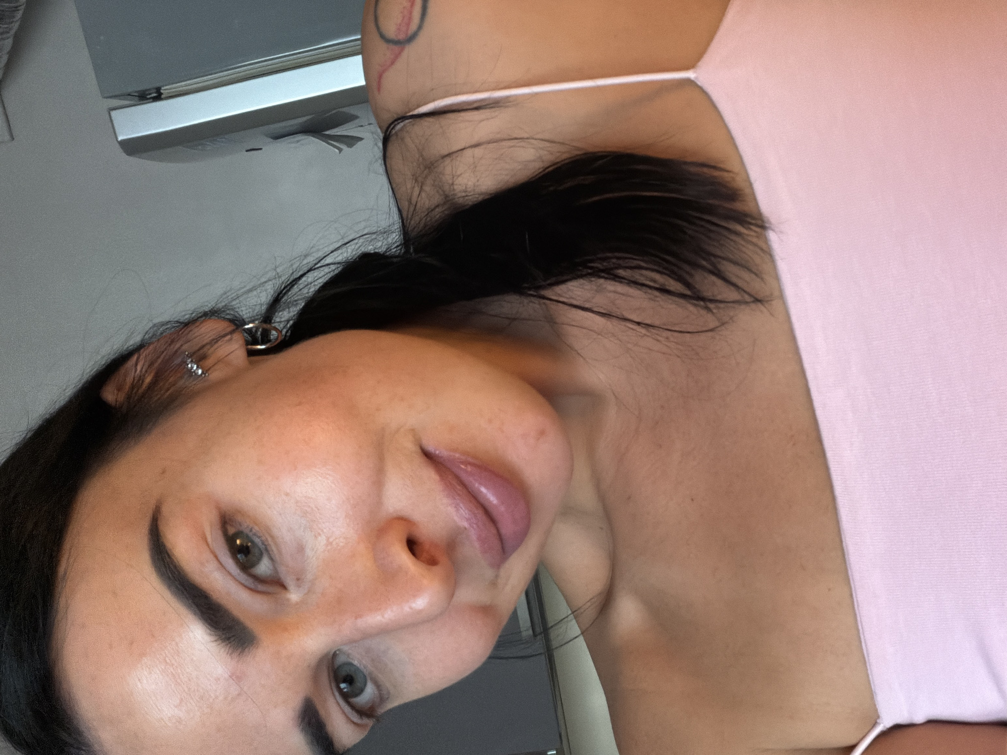 A close-up, slightly tilted shot depicts a woman with dark hair pulled back, fair skin, and light-colored eyes. She wears a light pink top with thin straps. Her eyebrows are dark and well-defined. Multiple piercings are visible on her ear. A tattoo is visible on her shoulder. The background is blurry and shows a metallic object and a gray wall, suggesting an indoor setting. No specific information indicates the location in Saint Petersburg, Russia.