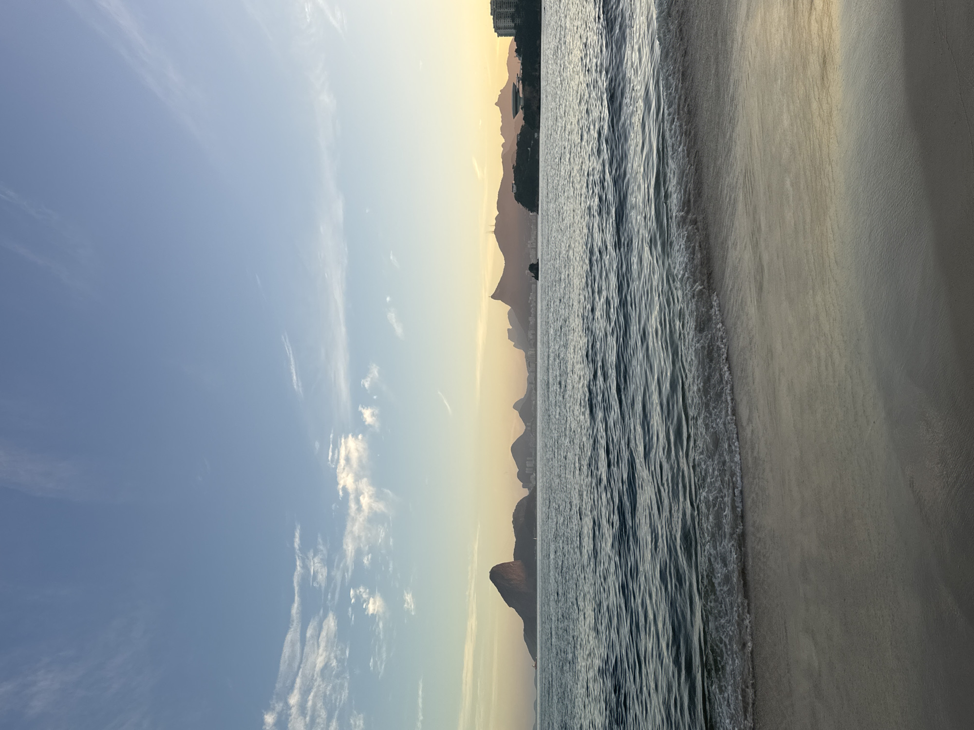 The image captures a serene coastal view of Niterói, Brazil, at either sunrise or sunset. In the foreground, a sandy beach is met by the gentle lapping of the ocean waves. The water is a deep blue, with white foam breaking near the shore. Beyond the water, the distinctive silhouette of Rio de Janeiro's iconic Sugarloaf Mountain and other mountains can be seen on the horizon. The sky is a gradient of pale blue at the top, transitioning to soft yellows and oranges near the horizon, indicating the time of day. A few wispy clouds dot the sky. In the middle ground, the city skyline of Rio de Janeiro is visible, with buildings and structures subtly illuminated by the warm light. No people are visible in the image, emphasizing the tranquility of the scene. The overall impression is one of natural beauty and peacefulness.