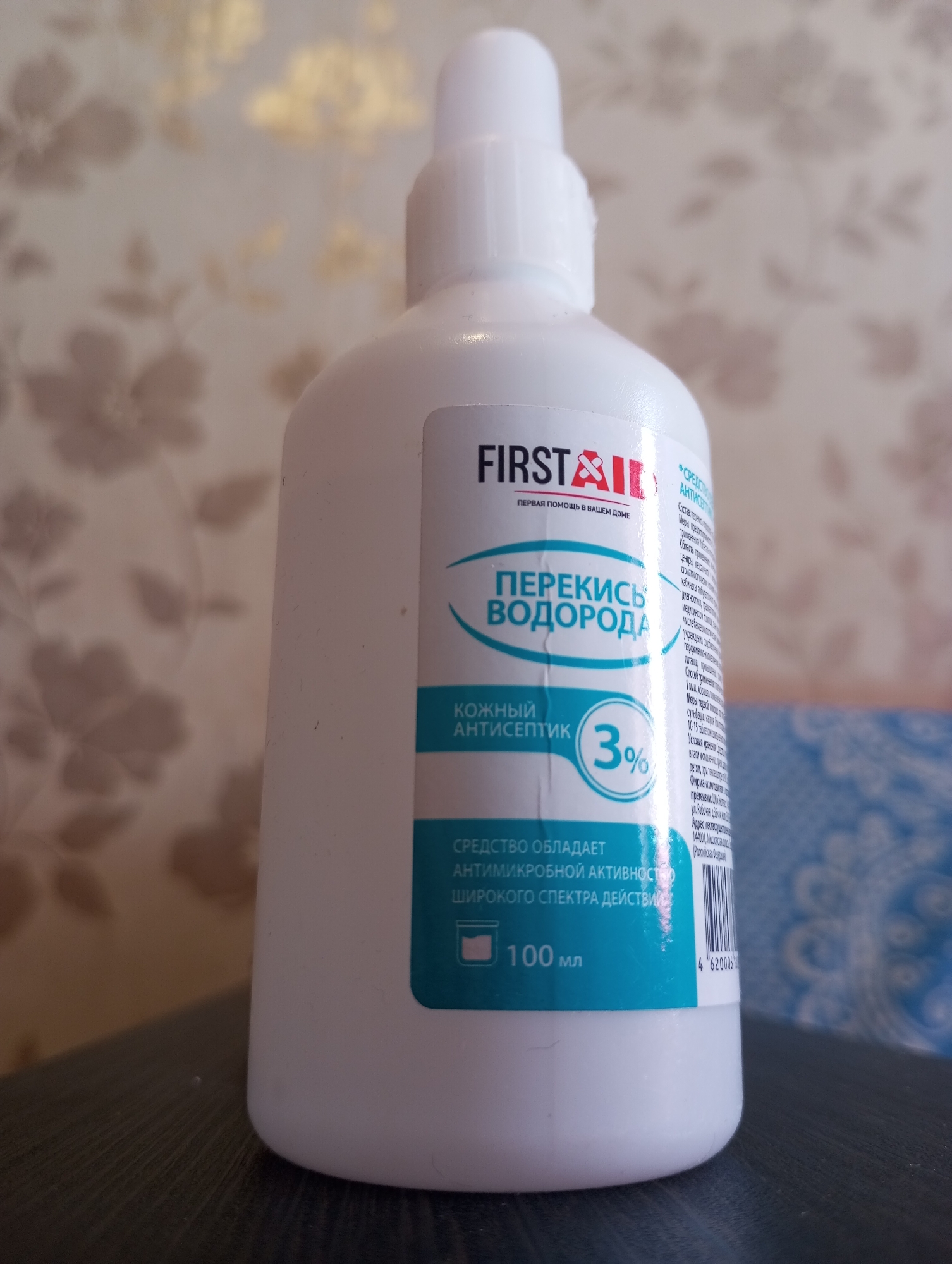 A close-up, slightly angled shot focuses on a white plastic bottle of "First Aid" hydrogen peroxide, 3% solution, for household use. The label is predominantly white with teal accents. The brand name "FIRST AID" is in black, with a red first aid cross logo. Below it, in smaller text, is "ПЕРВАЯ ПОМОЩЬ В ВАШЕМ ДОМЕ" (First aid in your home). The main text on the label reads "ПЕРЕКИСЬ ВОДОРОДА" (Hydrogen peroxide) in large teal lettering. Below this, "КОЖНЫЙ АНТИСЕПТИК" (Skin antiseptic) is written in smaller white text on a teal background, with "3%" in large numbers within a teal circle. The bottom of the label states "СРЕДСТВО ОБЛАДАЕТ АНТИМИКРОБНОЙ АКТИВНОСТЬЮ ШИРОКОГО СПЕКТРА ДЕЙСТВИЯ" (The product has broad-spectrum antimicrobial activity) and "100 мл" (100 ml). To the right of the main label, a smaller section of text details the composition and application, and a barcode with the number 4620006 is visible. The bottle is placed on a dark, possibly wooden surface, with a textured, light-colored, floral wallpaper in the background. The image does not contain any information about the city of Kirzhach, Russia.