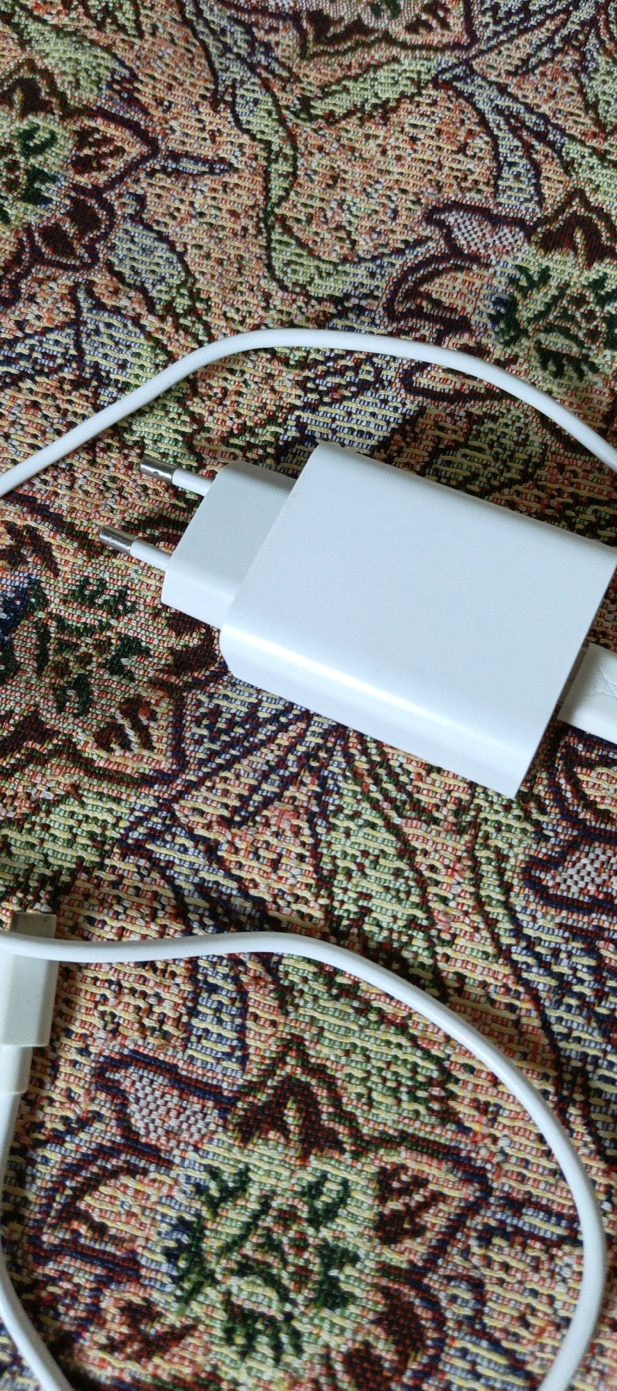 A white electrical charger and its white cable are displayed on a fabric surface with a colorful, intricate woven pattern. The charger is oriented horizontally, with its prongs visible. The cable extends from the charger and curves across the fabric. The background fabric exhibits a blend of red, green, yellow, and blue threads, forming a repeating abstract design. The scene is a close-up, tabletop view. There is no information in the image to suggest a location in Kot Sarang, Pakistan.
