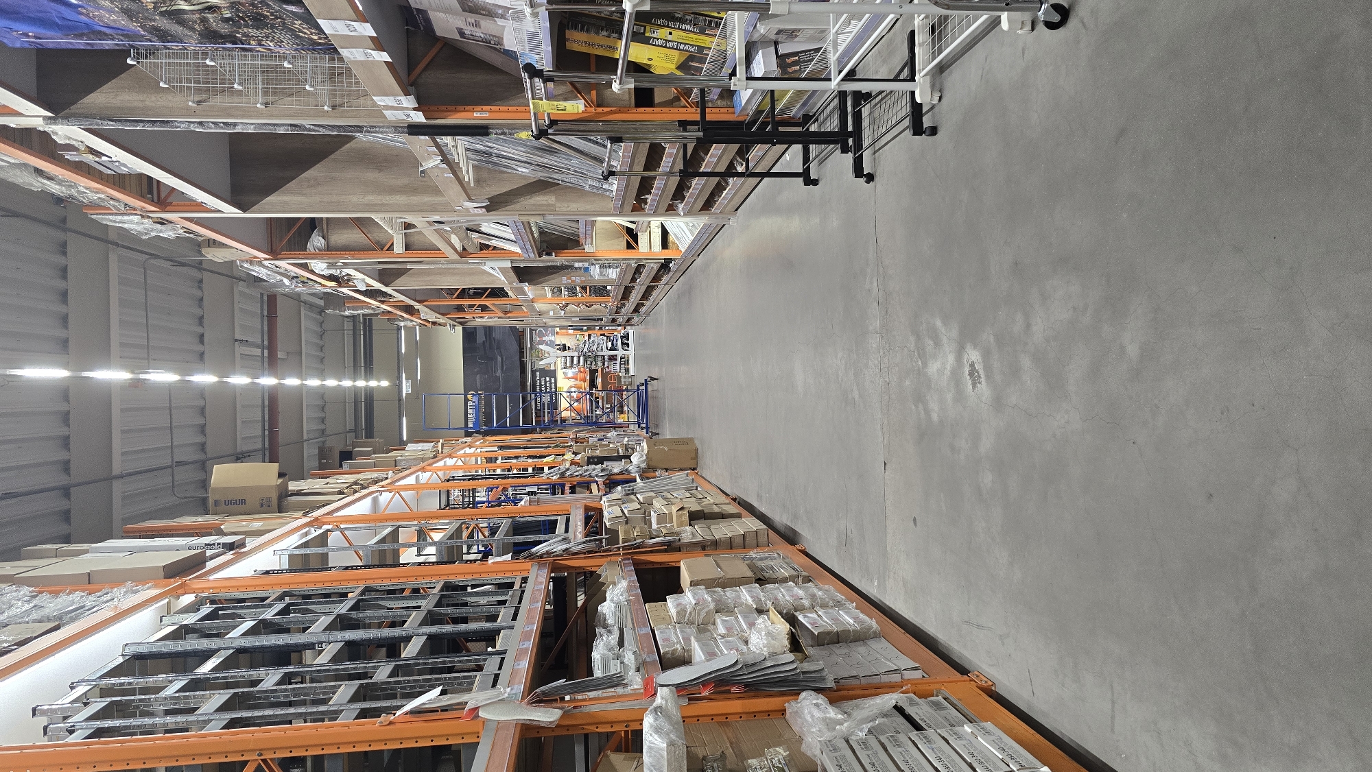 The image shows a long, indoor aisle in a warehouse or large retail store. The aisle is flanked by tall, orange metal shelving units filled with various goods, primarily packaged items and what appear to be building materials or hardware. The flooring is a smooth, gray concrete. In the distance, more shelving and some industrial-style carts are visible. The lighting comes from a series of bright, linear fluorescent fixtures running along the ceiling. The scene is clean and organized, suggesting a commercial setting. The image does not contain any people. The specific location in Zhytomyr, Ukraine is not ascertainable from the visual information present in the image.