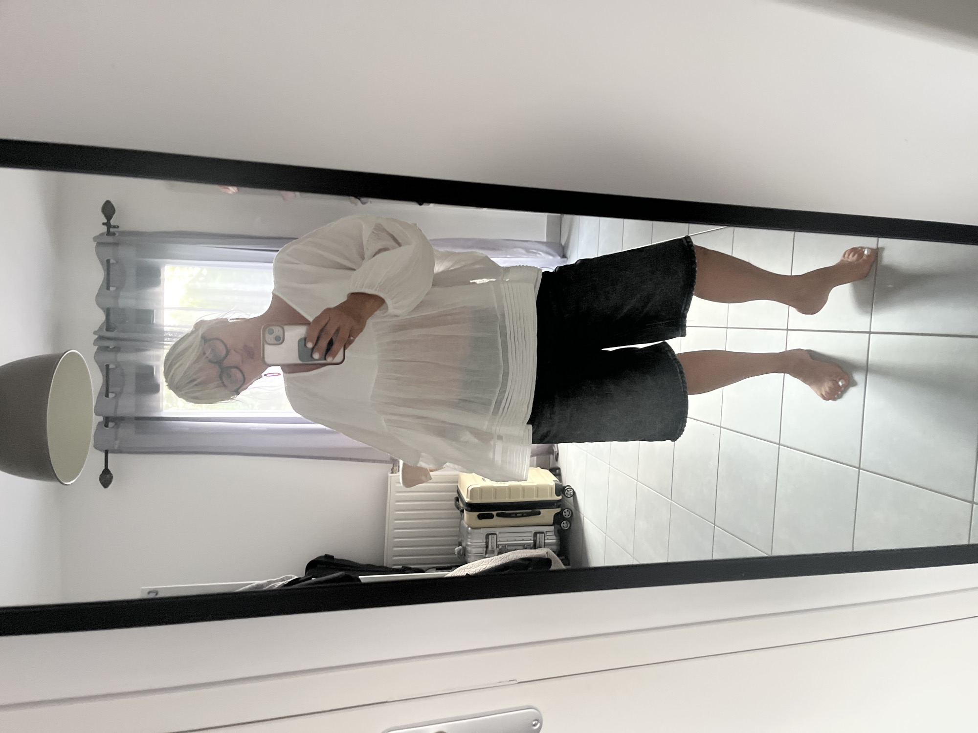 In a room with tiled flooring and light gray walls, a woman with blonde hair and glasses is reflected in a full-length mirror. She is wearing a loose white blouse with billowy sleeves and dark denim shorts. She is holding a smartphone in her right hand, as if taking a selfie. In the background, visible in the mirror, are two suitcases, a radiator, and gray curtains drawn across a window. The woman's feet are bare. The scene appears to be an indoor setting, possibly a bedroom or dressing area. The location is identified as Chambœuf, France.