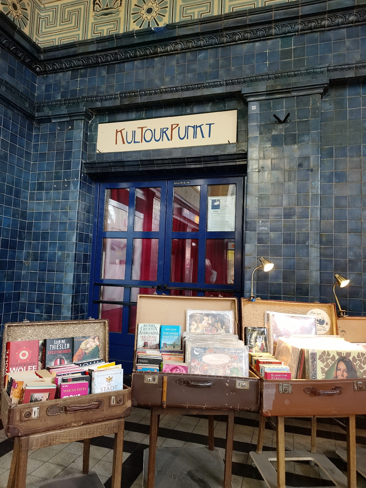 This image shows a shop selling books and records in Unknown, Unknown. The shop is called "Kultourpunkt" and is located in a building with blue tile walls. There are three suitcases filled with books and records on display. The suitcases are placed on top of small wooden tables. There are two small lamps clamped on the suitcase with records. There is a blue door behind the suitcases. The floor is tiled.
