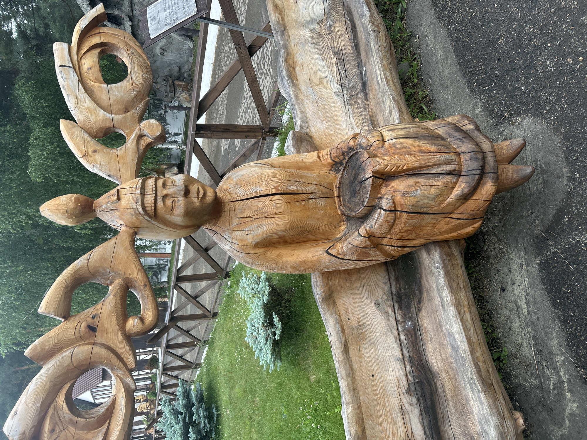 A weathered wooden sculpture of a seated figure with large, ornate antlers or headdress is positioned on a wooden bench in Savvateevka, Russia. The sculpture depicts a human-like face with closed eyes and a serene expression, wearing a conical hat. The figure's body is carved to represent clothing and it holds a wooden log in its lap. The bench and sculpture are made from rough-hewn logs, showing the natural grain and imperfections of the wood. Behind the sculpture, a wooden fence separates the area from a grassy lawn with small evergreen bushes. In the background, trees and a building with a brown roof are visible, along with a stone formation and a informational plaque. The ground in front of the bench appears to be paved with asphalt or gravel. The overall scene suggests an outdoor art installation or park setting.