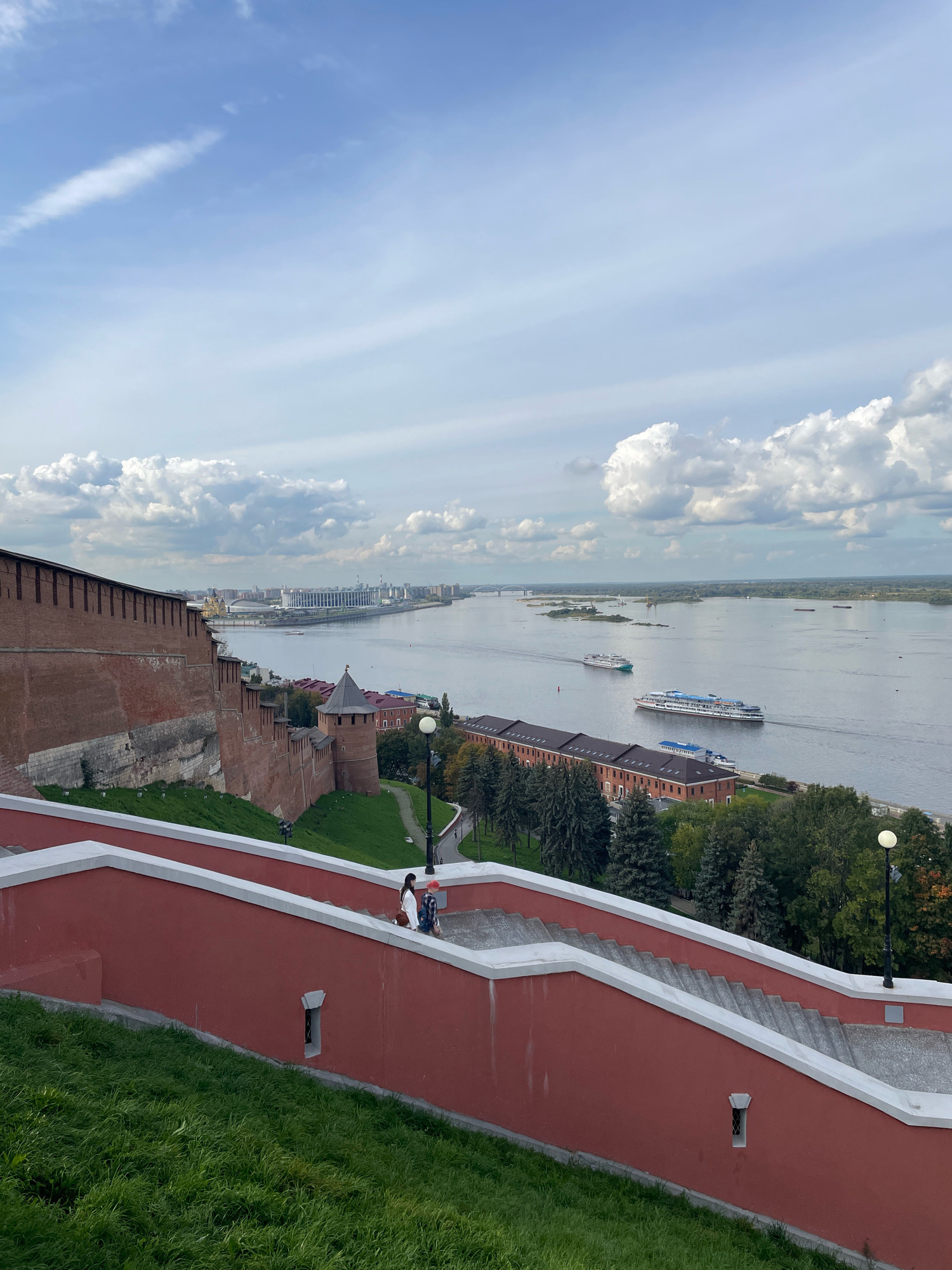 This image captures a scenic view of the Nizhny Novgorod Kremlin and the confluence of the Volga and Oka rivers. In the foreground, a red-walled staircase with white trim descends from the upper left to the lower right, with lush green grass covering the hillside. Two individuals, a woman with dark hair wearing a white top and dark pants, and a child in a red hat and blue checkered shirt, are ascending the stairs.
The middle ground reveals the imposing red brick walls of the Nizhny Novgorod Kremlin, a historic fortress with watchtowers and battlements. Beyond the Kremlin walls, the wide expanse of the Volga River stretches across the horizon. Several ships are visible on the river, including two large passenger vessels. The far bank of the river is lined with trees and buildings, with a modern stadium complex and bridges visible in the distance. The sky above is a mix of blue sky and fluffy white clouds, suggesting a pleasant day. The overall impression is one of historical grandeur and the beauty of the natural landscape.