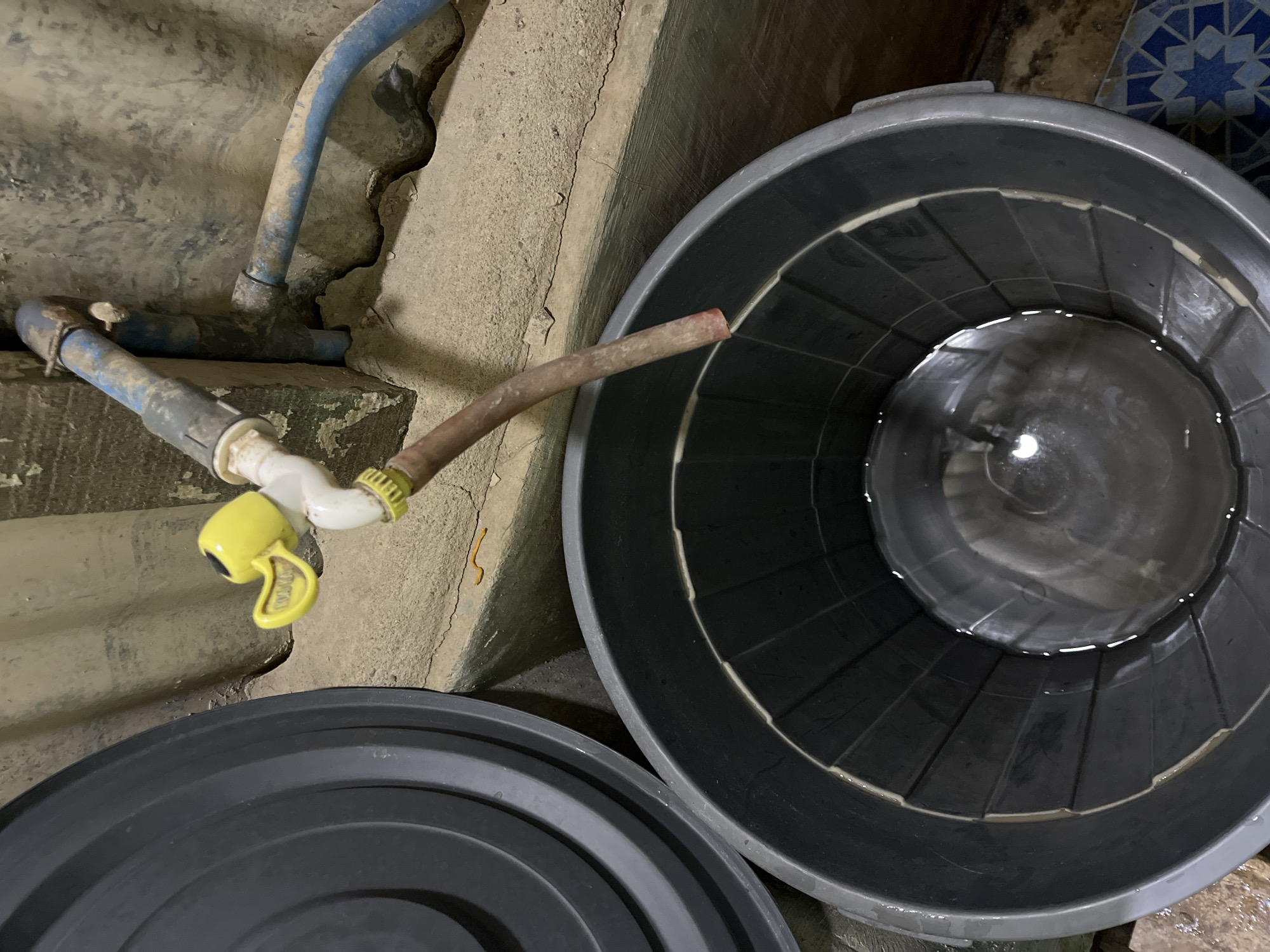 An overhead shot shows a large, dark gray plastic tub partially filled with clear water. The tub is positioned in front of a rough, concrete wall. To the left of the tub is the edge of another similar dark gray plastic container, its lid slightly ajar. Above and to the right of the tub, a spigot with a yellow duck-shaped handle is attached to plumbing pipes. A brown hose extends from the spigot and hangs over the edge of the tub, dripping water into it. The environment appears to be utilitarian, possibly an outdoor or semi-outdoor utility area. The location is specified as Benuaraja, Indonesia.