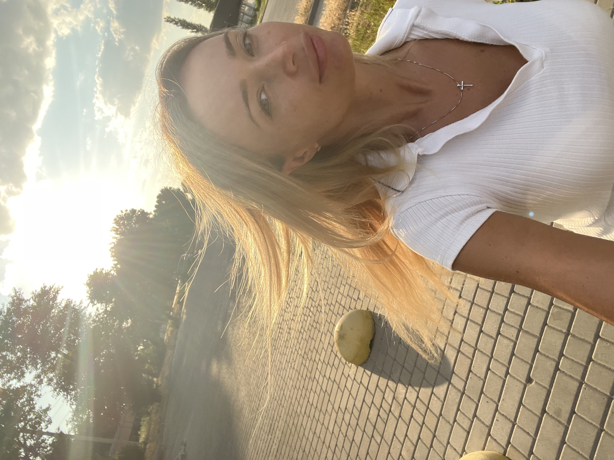 The image shows a woman in her late 30s or early 40s taking a selfie outdoors. She has blonde hair and is wearing a white polo shirt. A silver necklace with a cross pendant is visible. The background suggests an urban setting with a paved walkway, some trees, and a bright sky with clouds. The sun is low in the sky, casting long shadows and creating a lens flare effect. There is a round, yellow object on the walkway in the lower left portion of the image. The scene appears to be in Fontanka, Ukraine, based on the provided information.