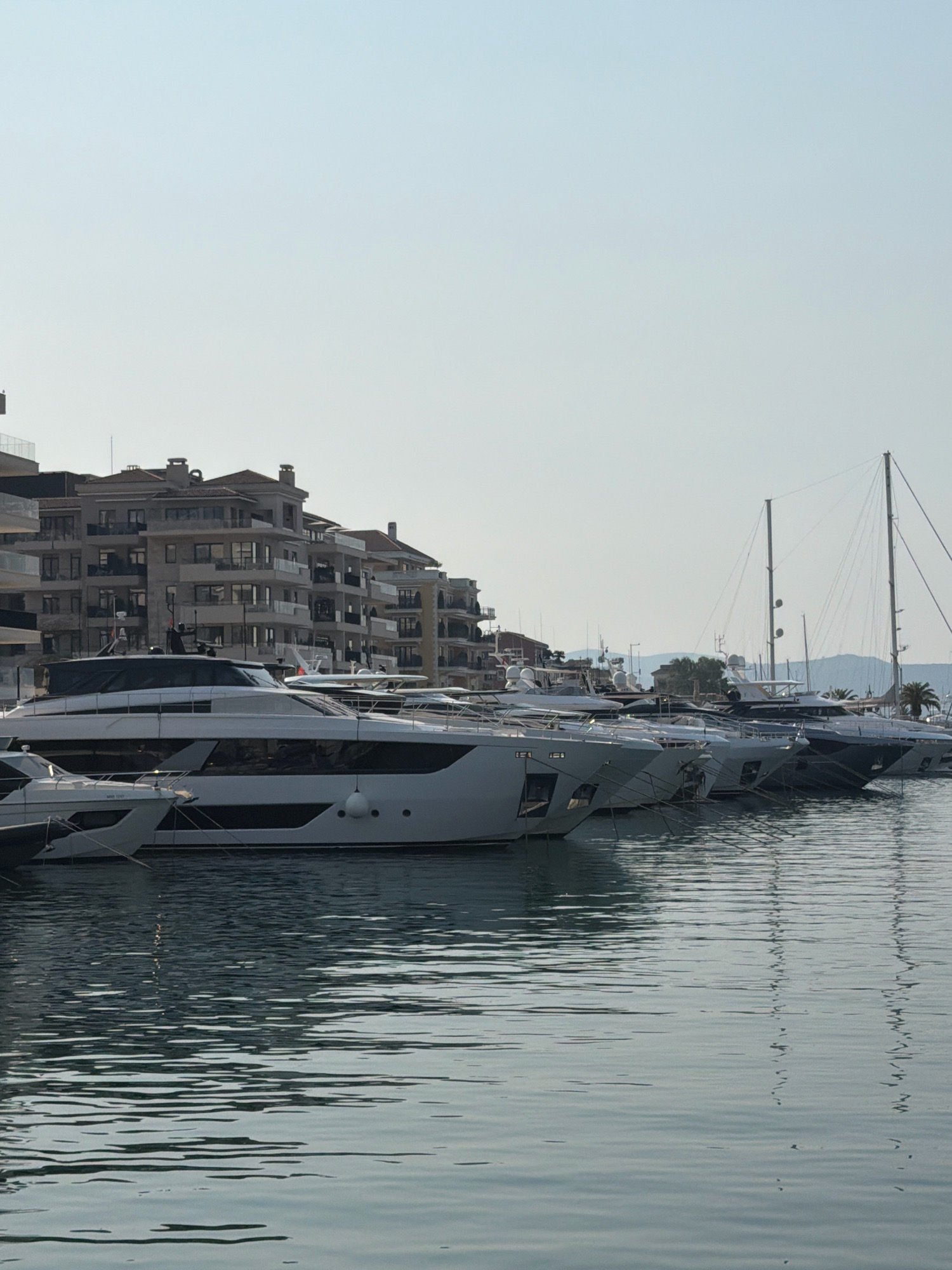 The image captures a serene harbor scene in Tivat, Montenegro. Several modern yachts are docked in the foreground, their sleek white hulls reflecting in the calm, rippling water. Behind the yachts, a multi-story building with a tan facade and numerous windows lines the waterfront, suggesting a residential or hotel complex. The sky is clear and pale blue, indicating a bright, possibly early morning or late afternoon. The overall impression is one of luxury and tranquility.