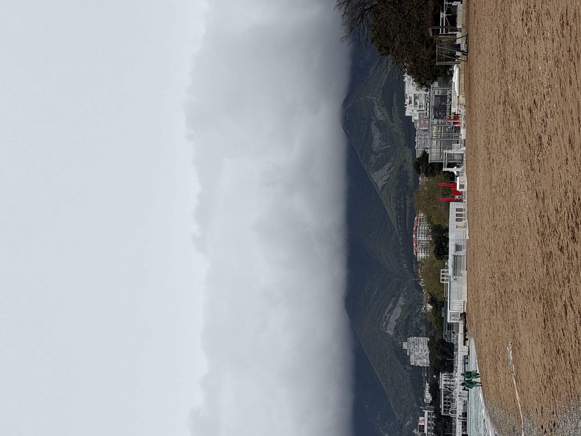 The image depicts a landscape featuring a beach, mountains shrouded in fog, and buildings. The beach is sandy and extends into the foreground. Beyond the beach, a row of buildings is visible, appearing as white structures with red accents. These buildings seem to be located at the foot of a mountain range. The mountains are partially obscured by thick fog, giving the scene a misty and somewhat dramatic atmosphere. The sky is overcast and blends seamlessly with the fog. The image is rotated ninety degrees clockwise.