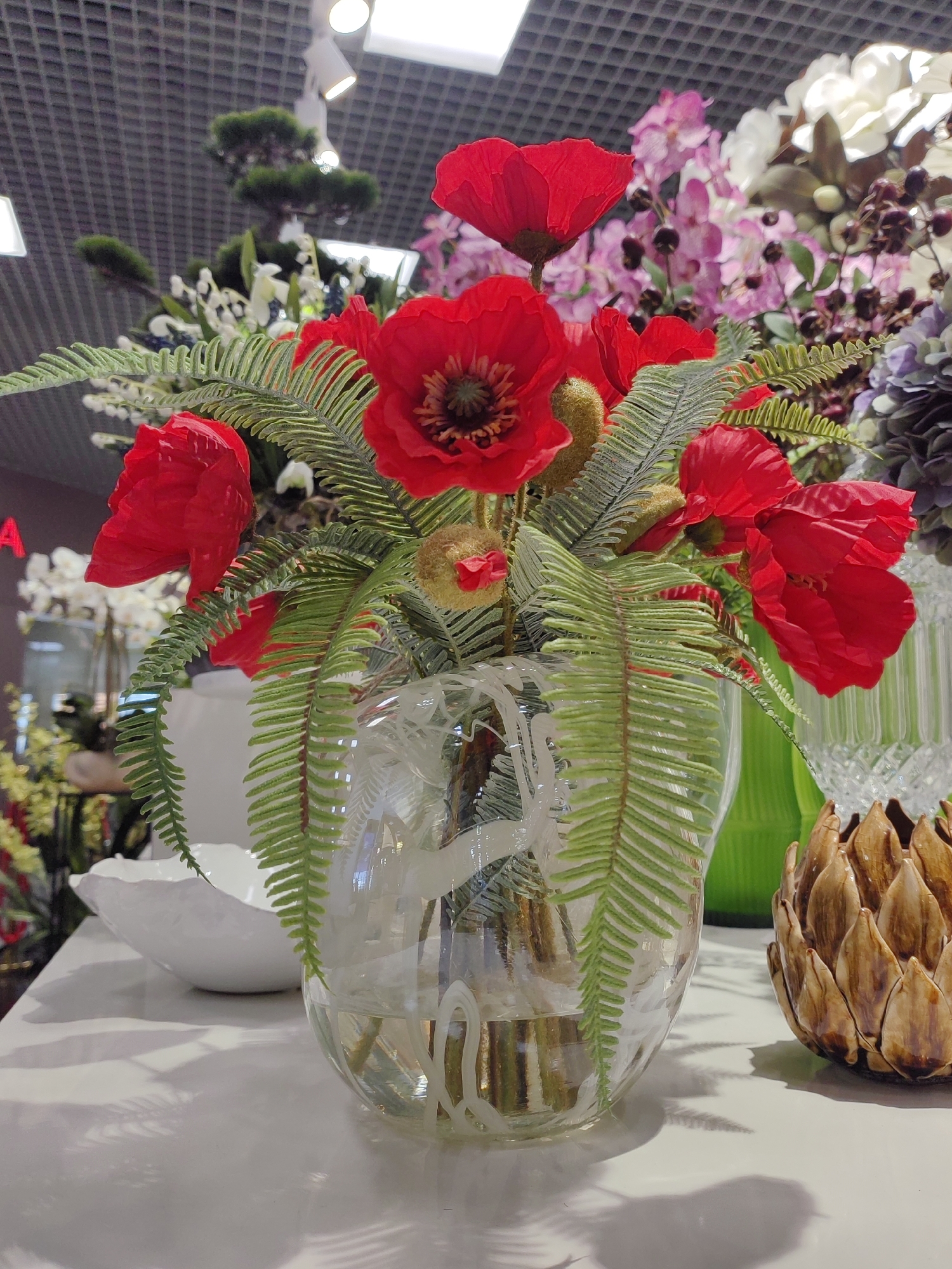 This media file depicts a still life arrangement of artificial flowers and decorative objects on a white surface, likely a tabletop or display counter. The central subject is a clear glass vase filled with water and containing a bouquet of vibrant red poppies and lush green fern fronds. The poppies have delicate, crinkled petals and dark centers, characteristic of the flower. The fern leaves are finely dissected and add a dramatic contrast to the bold red of the poppies.
Behind the main floral arrangement, a variety of other artificial flowers are visible, including clusters of pink orchids and white blossoms, along with darker, berry-like accents and muted purple foliage, creating a rich, textured background.
To the right of the vase, a ceramic decorative object resembling an artichoke or a pinecone, with a textured, layered surface in earthy brown and gold tones, sits prominently. To the left and slightly behind the vase, a large, irregular white ceramic bowl is partially visible. The background suggests an indoor setting, possibly a home decor store or a showroom, with a textured ceiling and recessed lighting fixtures visible at the top of the image. The overall impression is one of decorative elegance, with a focus on natural forms rendered in artificial materials. There are no people or animals visible, and the scene is static, conveying a sense of display rather than action. The lighting appears to be artificial, casting soft shadows on the white surface. No discernible text is visible within the image. The context of Saint Petersburg, Russia, is provided, but no specific visual cues within the image directly confirm this location.